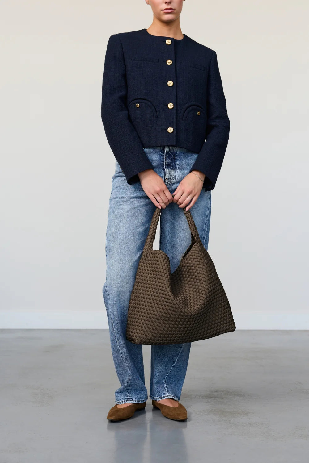 Person wearing a navy jacket and blue jeans, holding a brown woven handbag against a plain background.