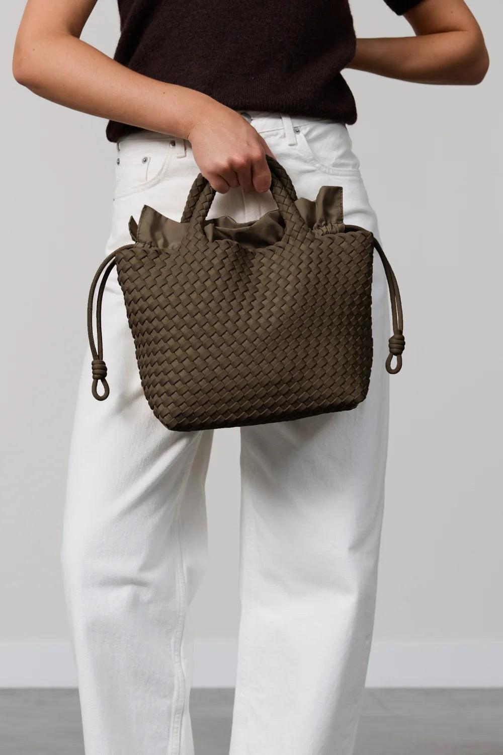 A person wearing white pants and a dark brown top holds a small woven brown handbag with top handles and drawstring details against a plain light gray studio background.