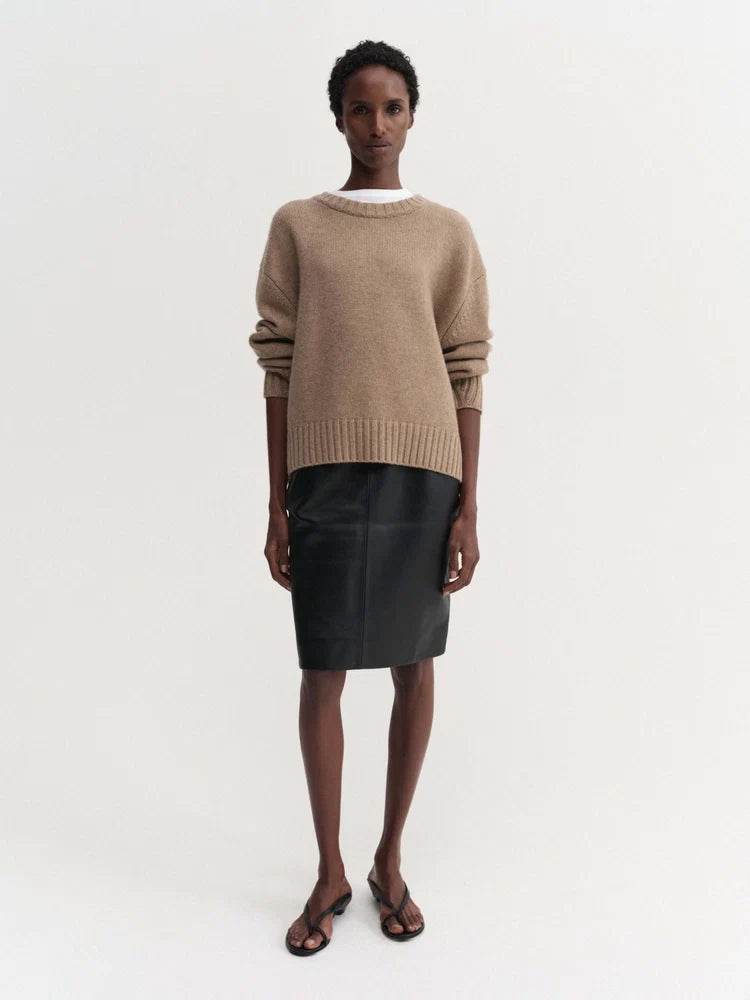 A model stands against a plain light background wearing a beige knit sweater, a black leather pencil skirt, and dark brown flat sandals.