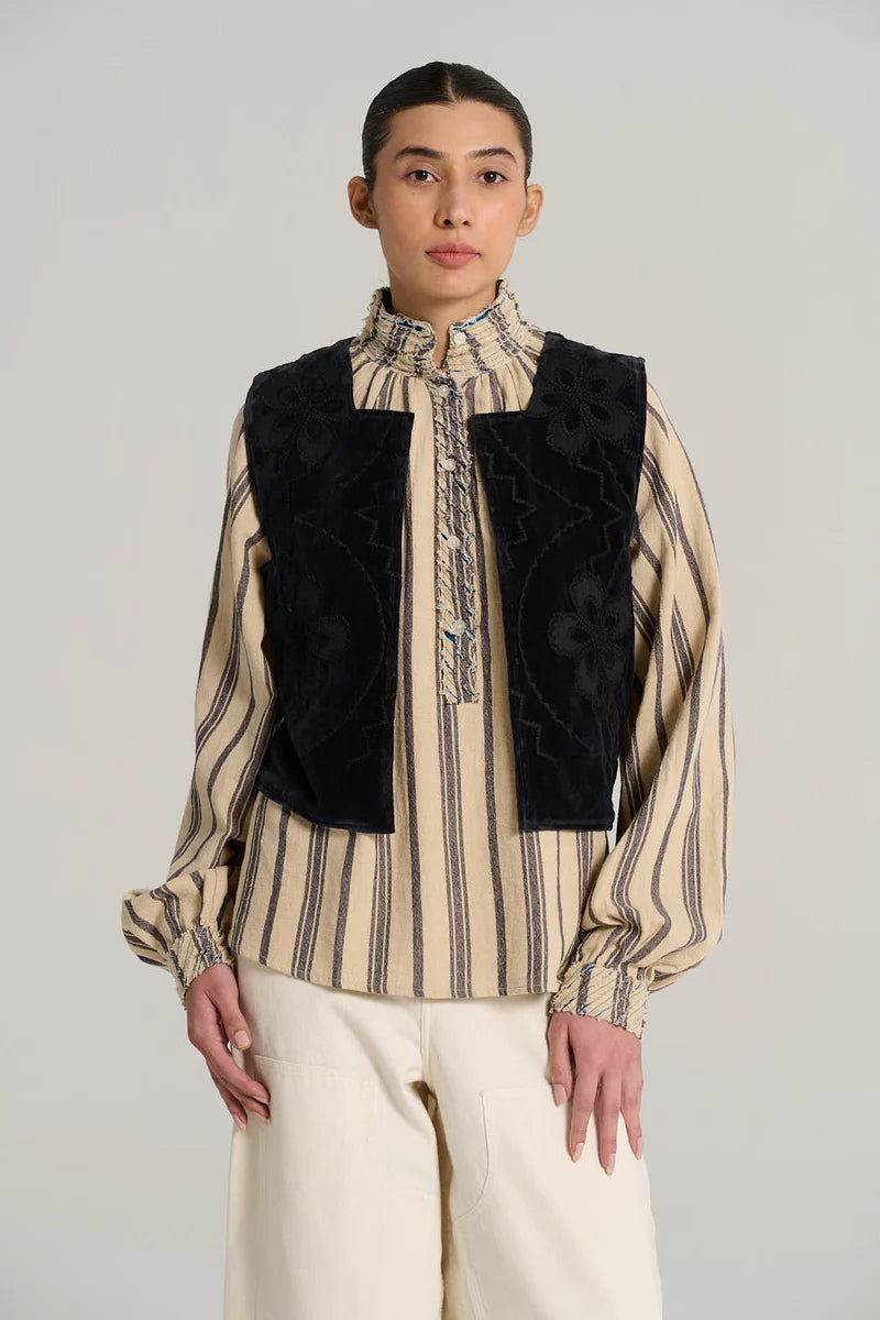 A model wearing a cream and navy vertically striped long-sleeve blouse with a high ruffled collar, paired with a black embroidered vest and cream trousers, against a light gray studio background.