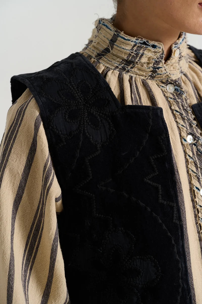 Close-up of a person wearing a black embroidered vest over a beige and black striped high-neck blouse.