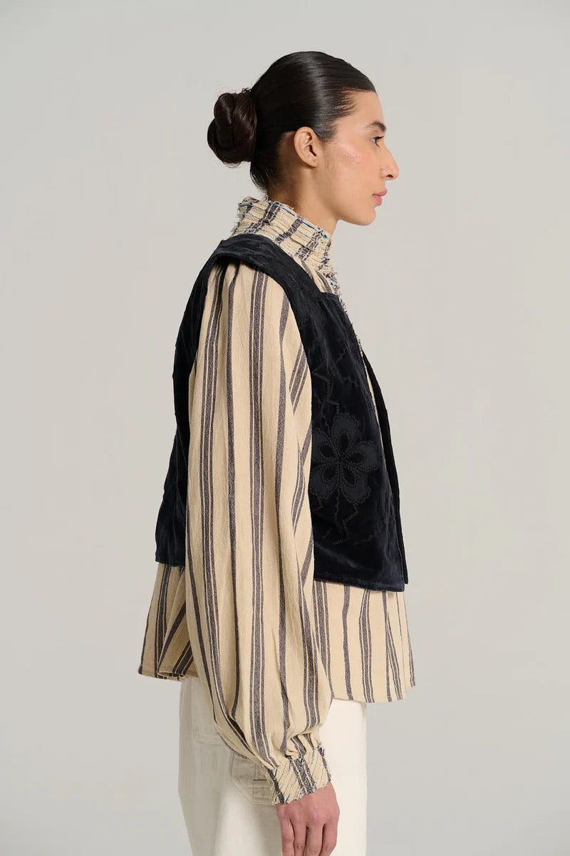 A model shown in profile wearing a dark navy velvet vest over a beige and navy vertically striped long-sleeve blouse, paired with cream trousers, against a light gray studio background.