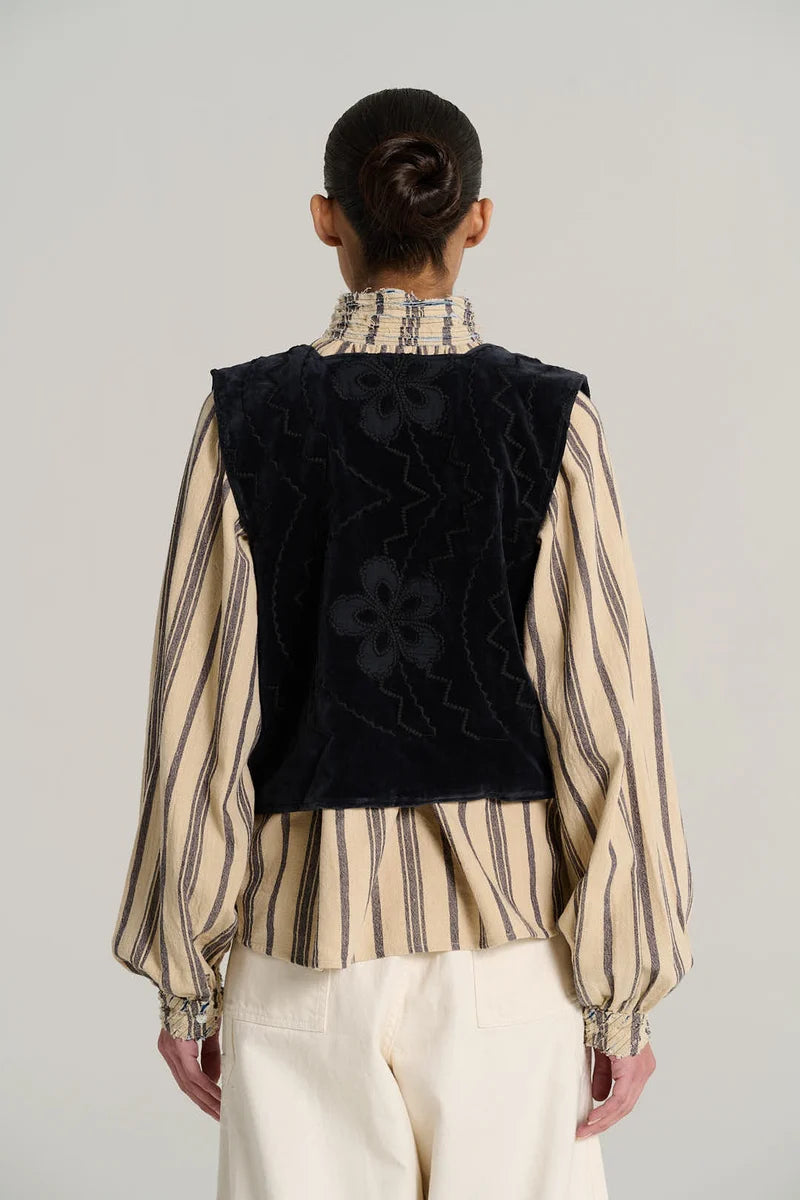 A model shown from the back wearing a cream and brown vertically striped long-sleeve blouse with a high ruffled collar, a black velvet vest with tonal floral embroidery, and cream trousers, against a light gray studio background.