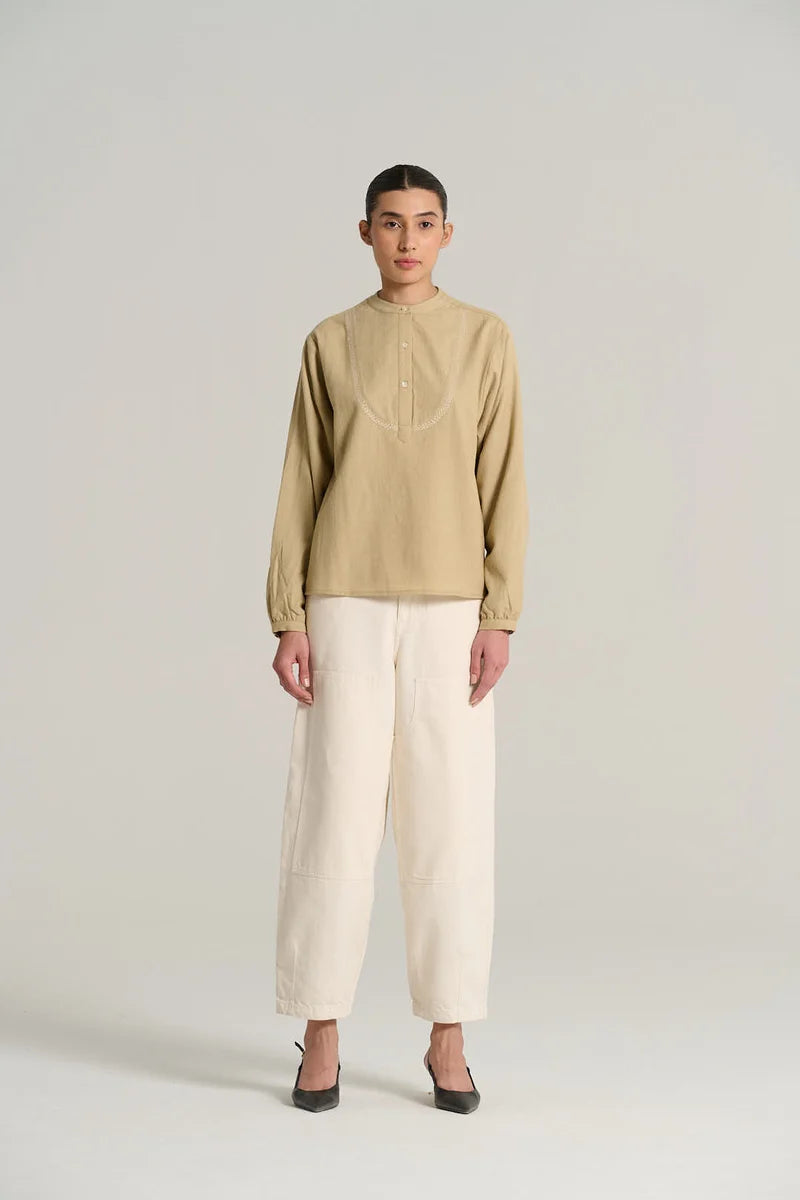 A model stands against a light gray studio background wearing a beige long-sleeve mock-neck top with tonal embroidery, paired with off-white wide-leg trousers and black pointed shoes.