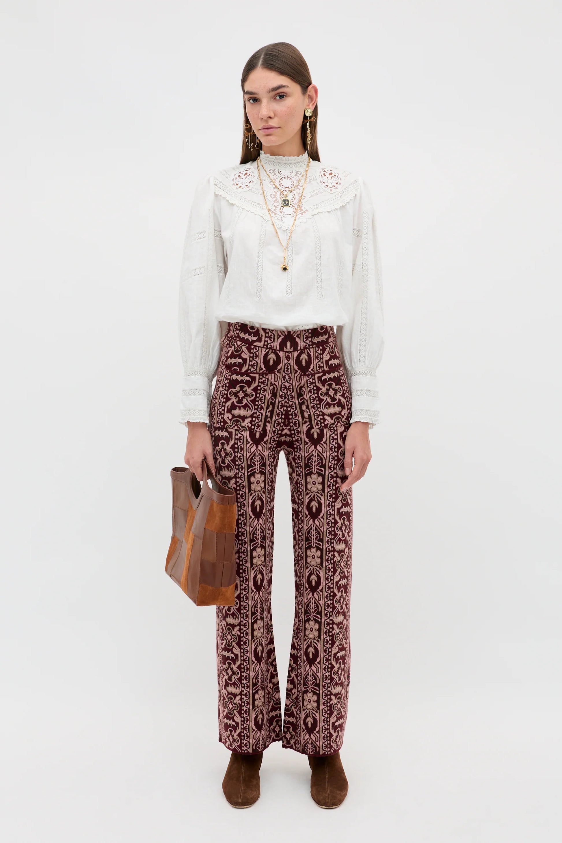 A model stands against a light gray studio background wearing a white long-sleeve blouse with a decorative lace-like collar, high-waisted patterned trousers in burgundy and beige, brown ankle boots, and holding a brown leather tote bag.
