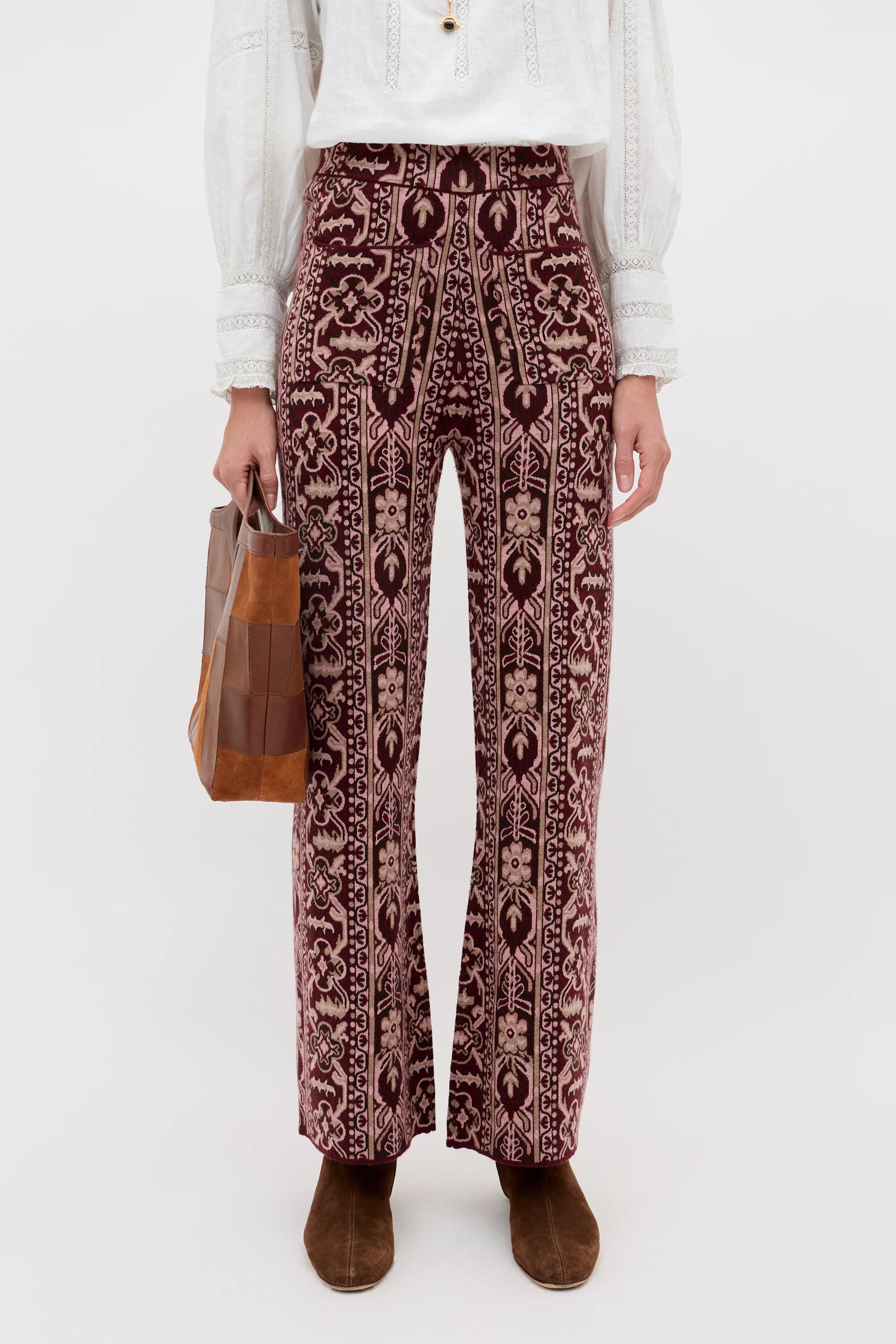 A model is shown from the waist down wearing high-waisted, wide-leg trousers with a dark burgundy and beige bohemian pattern. The model is also wearing a white long-sleeve blouse and brown suede boots, and holding a brown leather tote bag against a plain white studio background.
