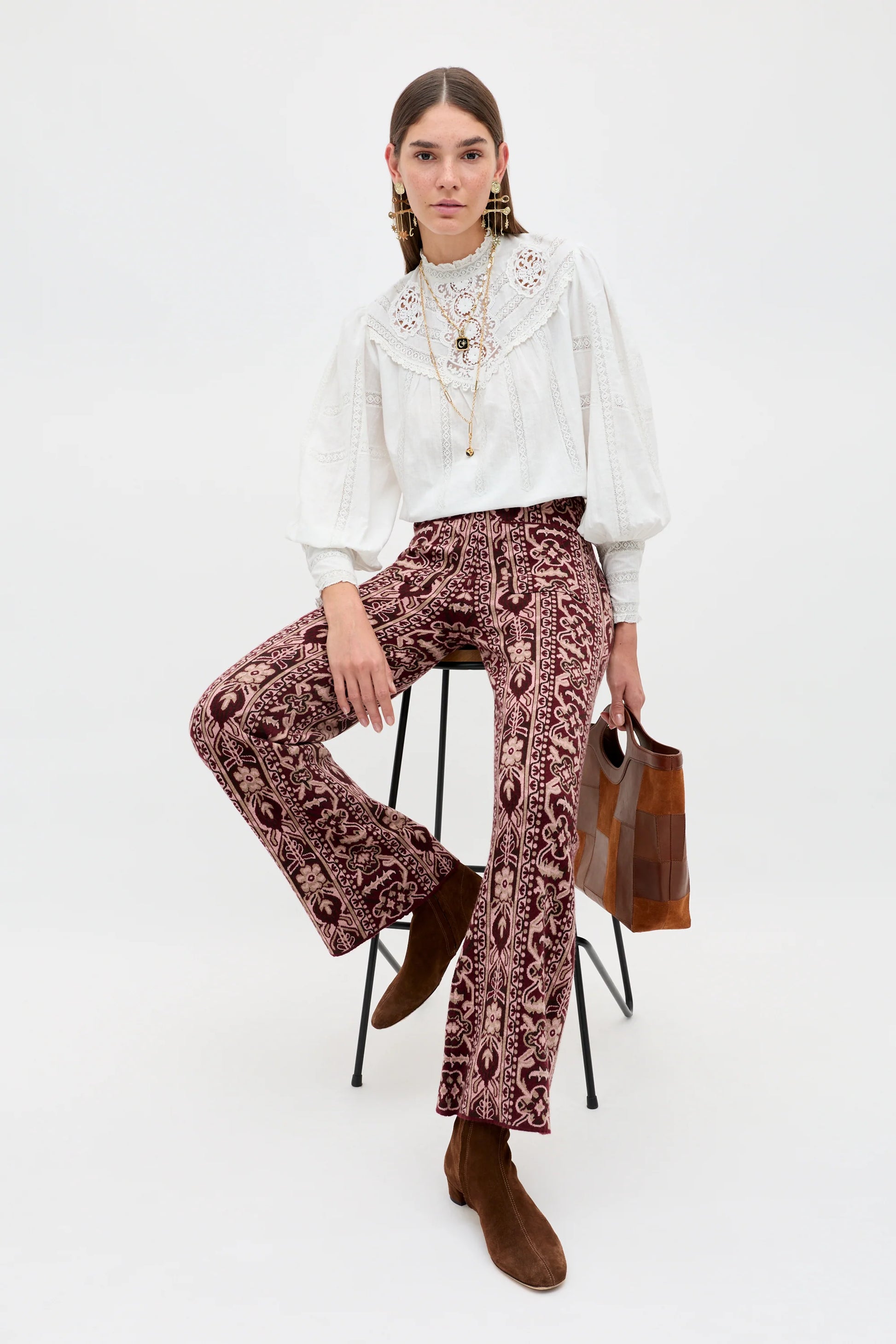 A model sits on a black metal stool wearing a white long-sleeve blouse with a ruffled collar, brown patterned wide-leg trousers, and brown suede boots, holding a brown leather handbag against a plain white studio background.
