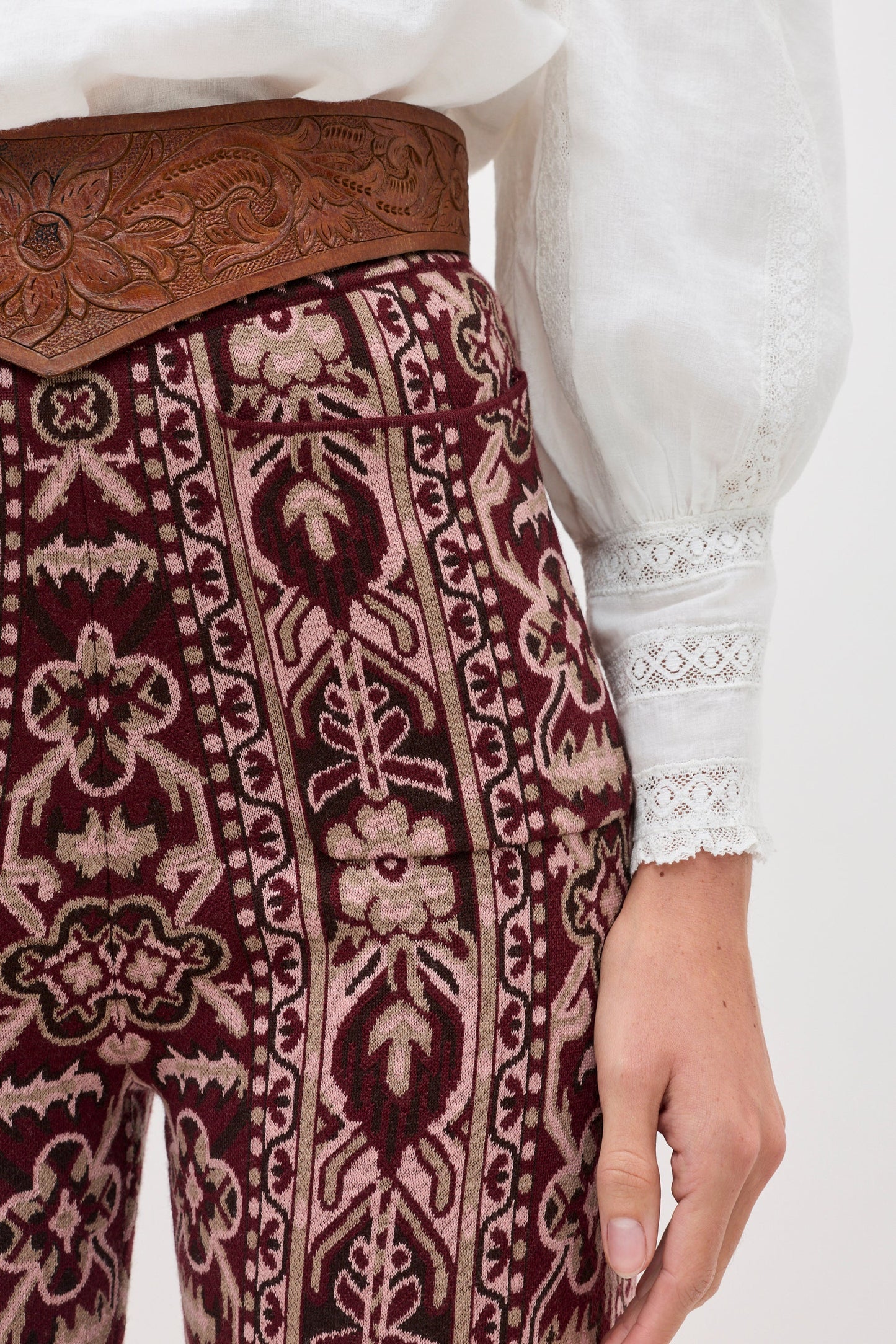 A close-up of a person wearing patterned dark red and beige woven pants with a brown tooled leather belt, a white lace-trimmed blouse, and a light gray background.