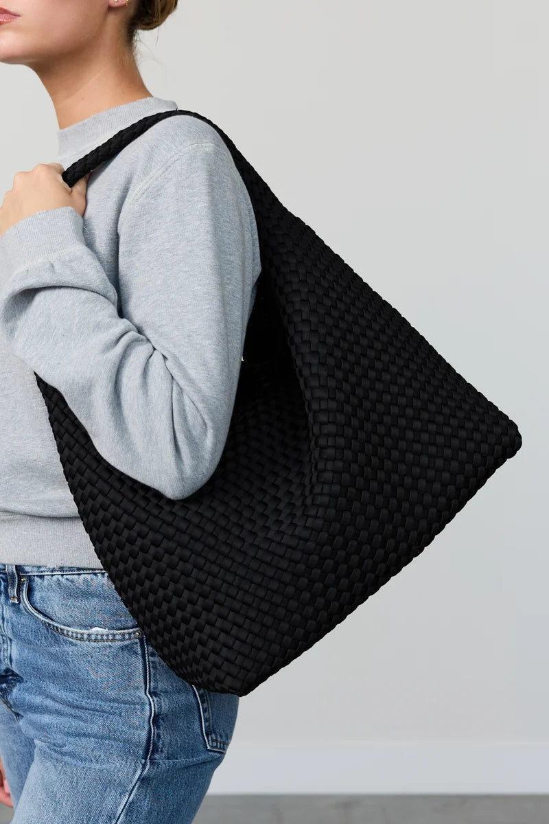 A person wearing a light gray sweatshirt and blue jeans carries a large black woven tote bag over their shoulder against a plain light gray wall.