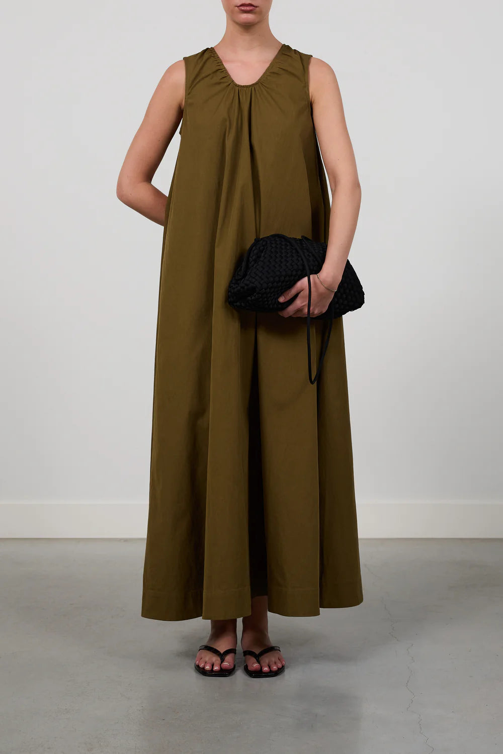 A model wearing a sleeveless, floor-length olive green dress, holding a black textured clutch bag, standing against a neutral light gray wall.