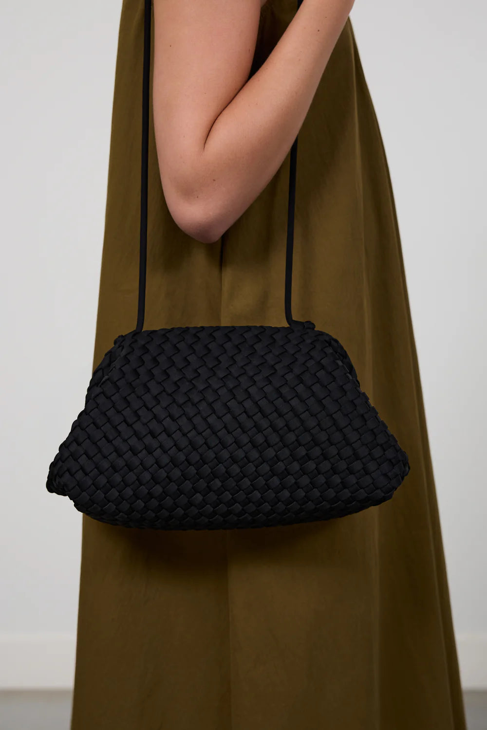A black woven shoulder bag with a thin strap, worn crossbody over an olive green dress against a plain light gray background.