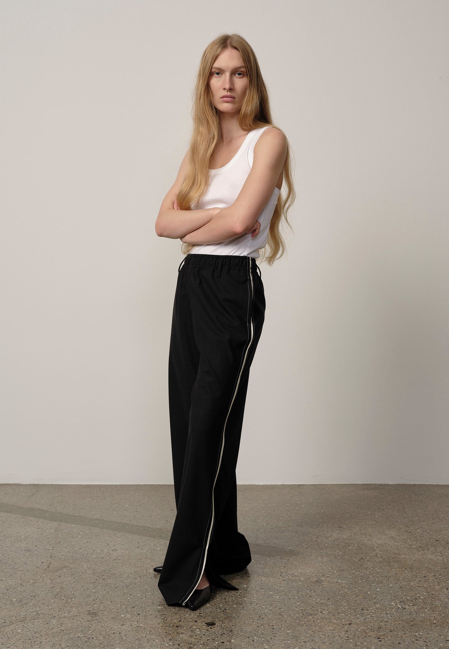 Woman wearing a white sleeveless top and black pants with a white stripe against a plain background