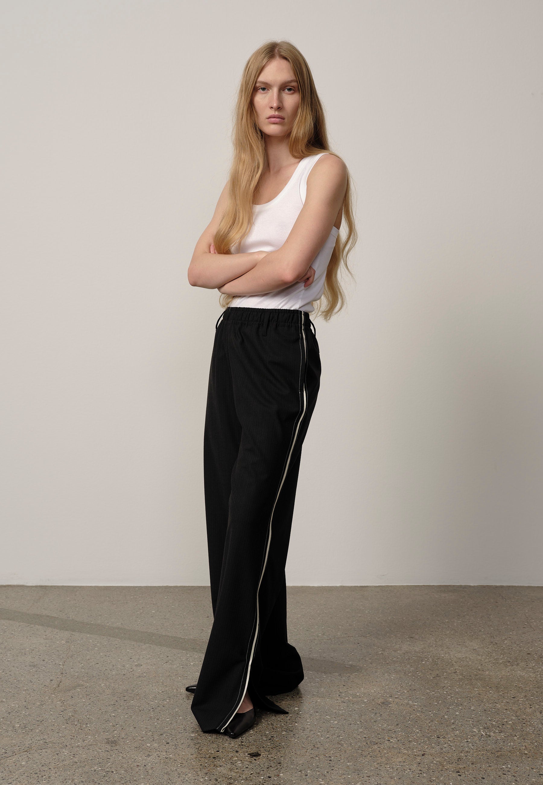 Woman wearing a white sleeveless top and black pants with a white stripe against a plain background
