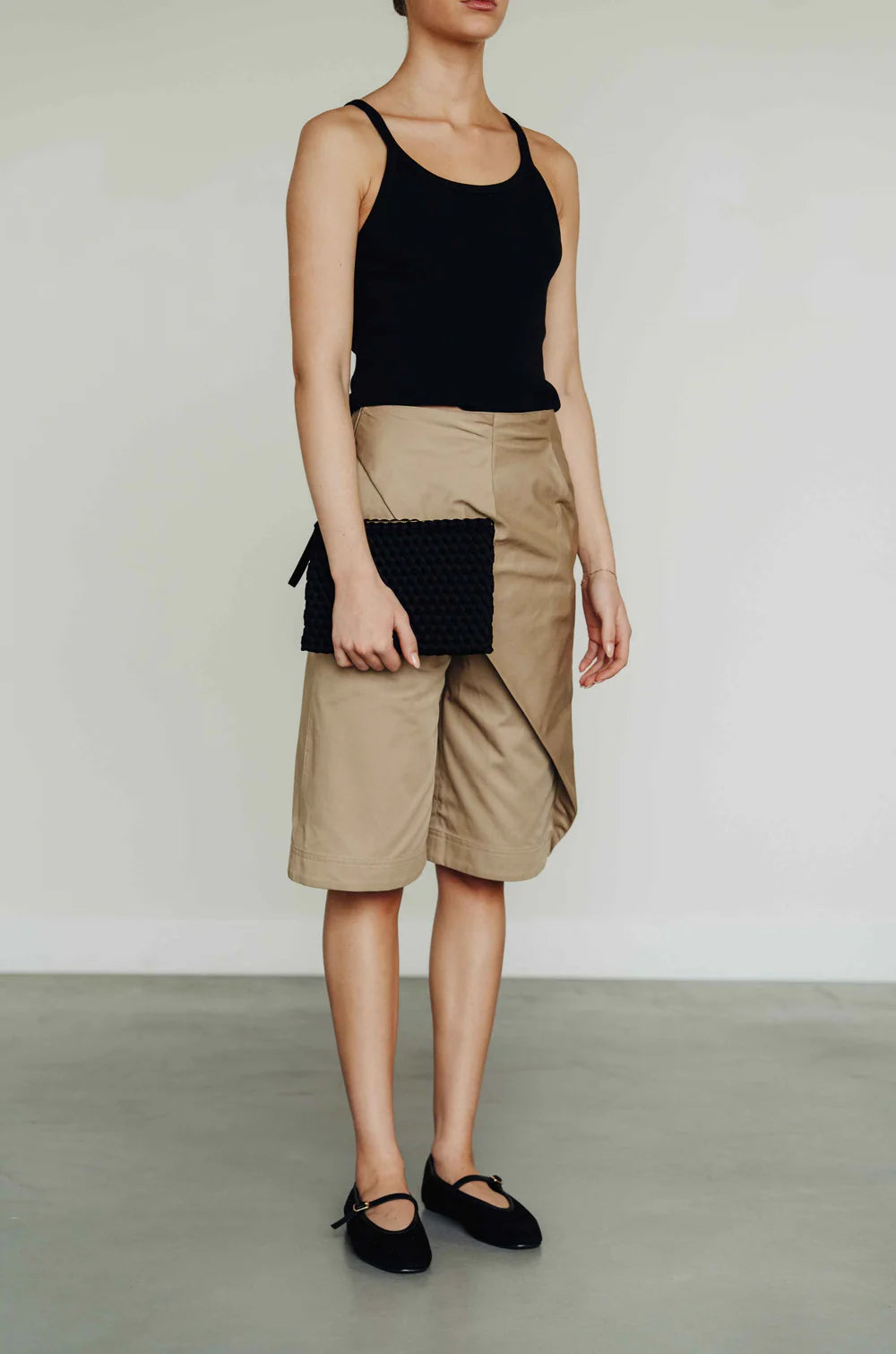 A model wearing a black ribbed tank top, beige wrap-style knee shorts, and black ballet flats, holding a black woven clutch bag against a plain light gray studio background.