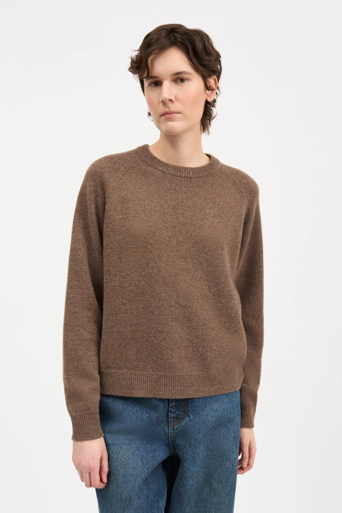 A model wearing a brown crewneck knit sweater and blue jeans, standing against a plain white studio background.