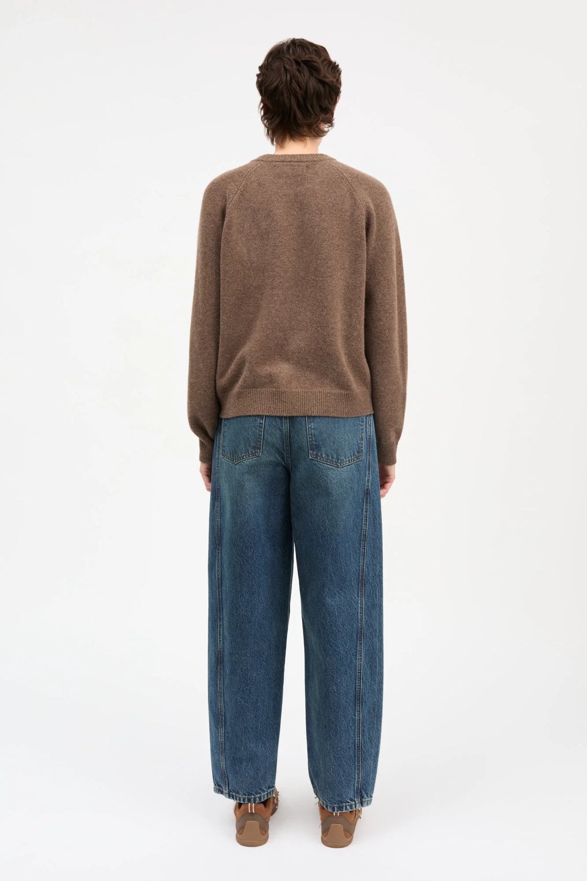 A person shown from the back wearing a brown knit sweater and wide-leg blue jeans, standing against a plain light gray studio background.