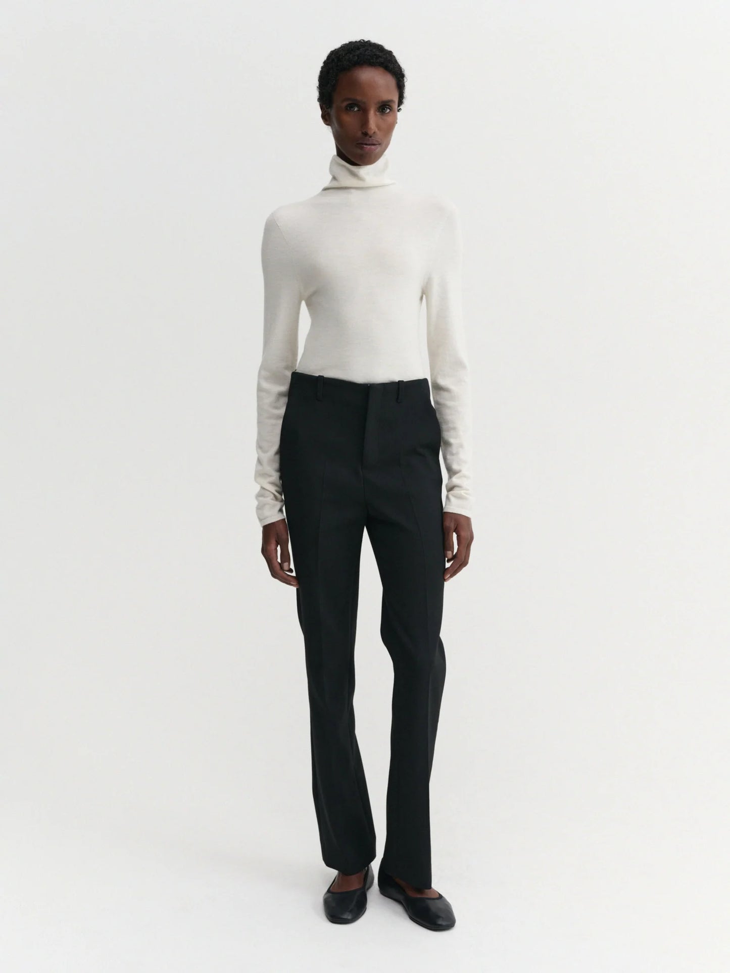 A model stands against a plain white studio background wearing a fitted white turtleneck sweater, black trousers, and dark flat shoes.
