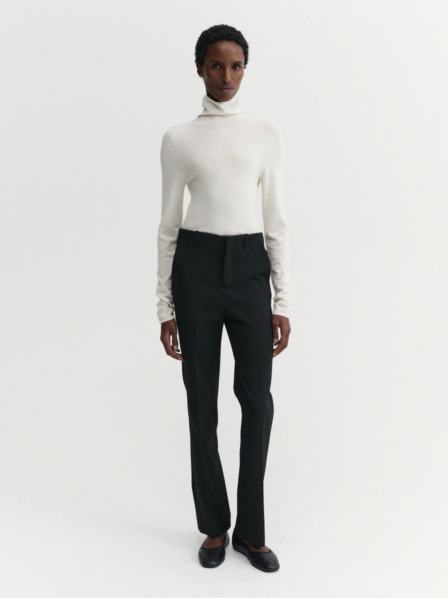 A model stands against a plain white studio background wearing a fitted white turtleneck sweater, black trousers, and dark flat shoes.