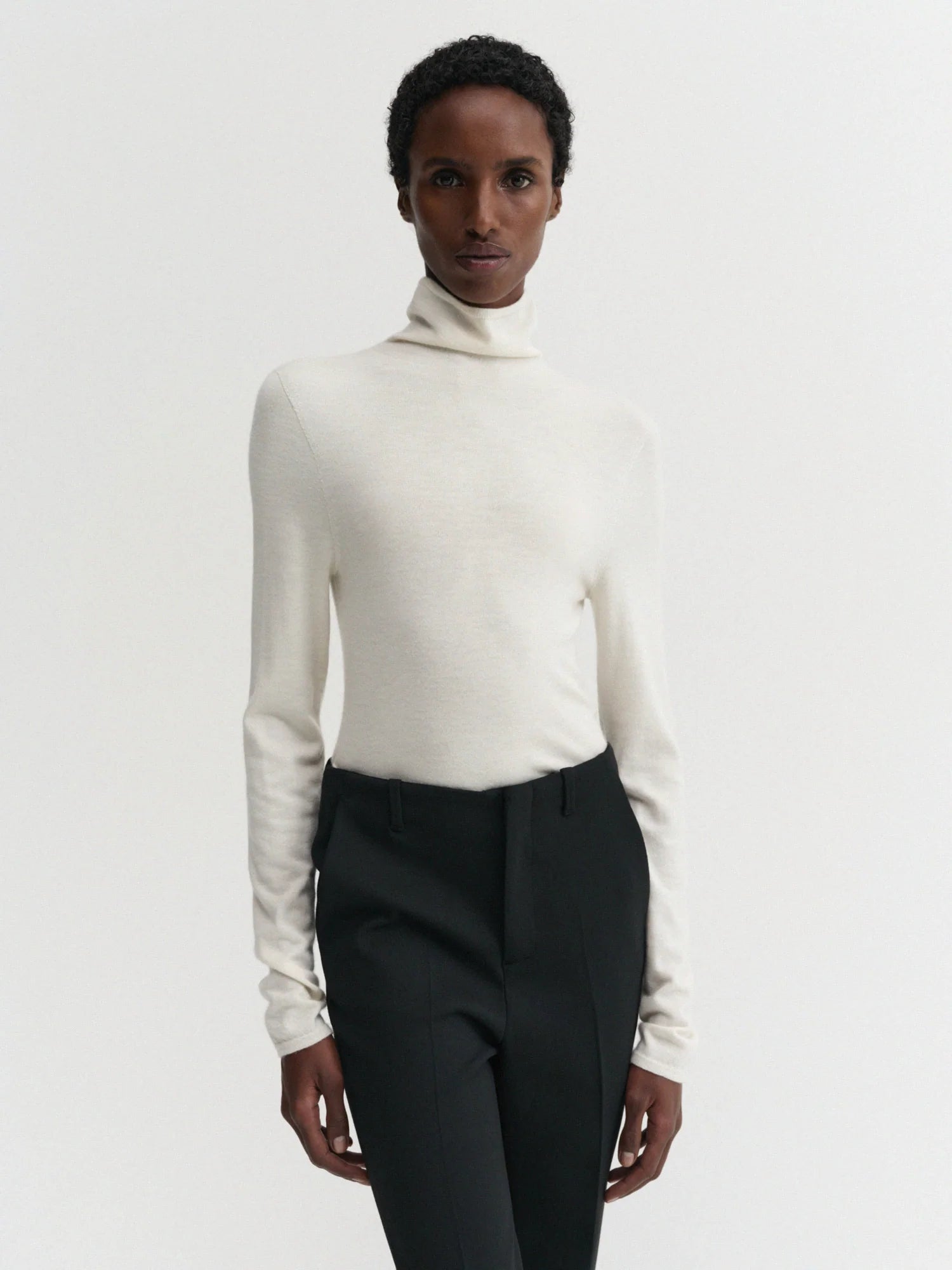 A model wearing a cream-colored, long-sleeve turtleneck sweater paired with black trousers, photographed against a solid light gray background.