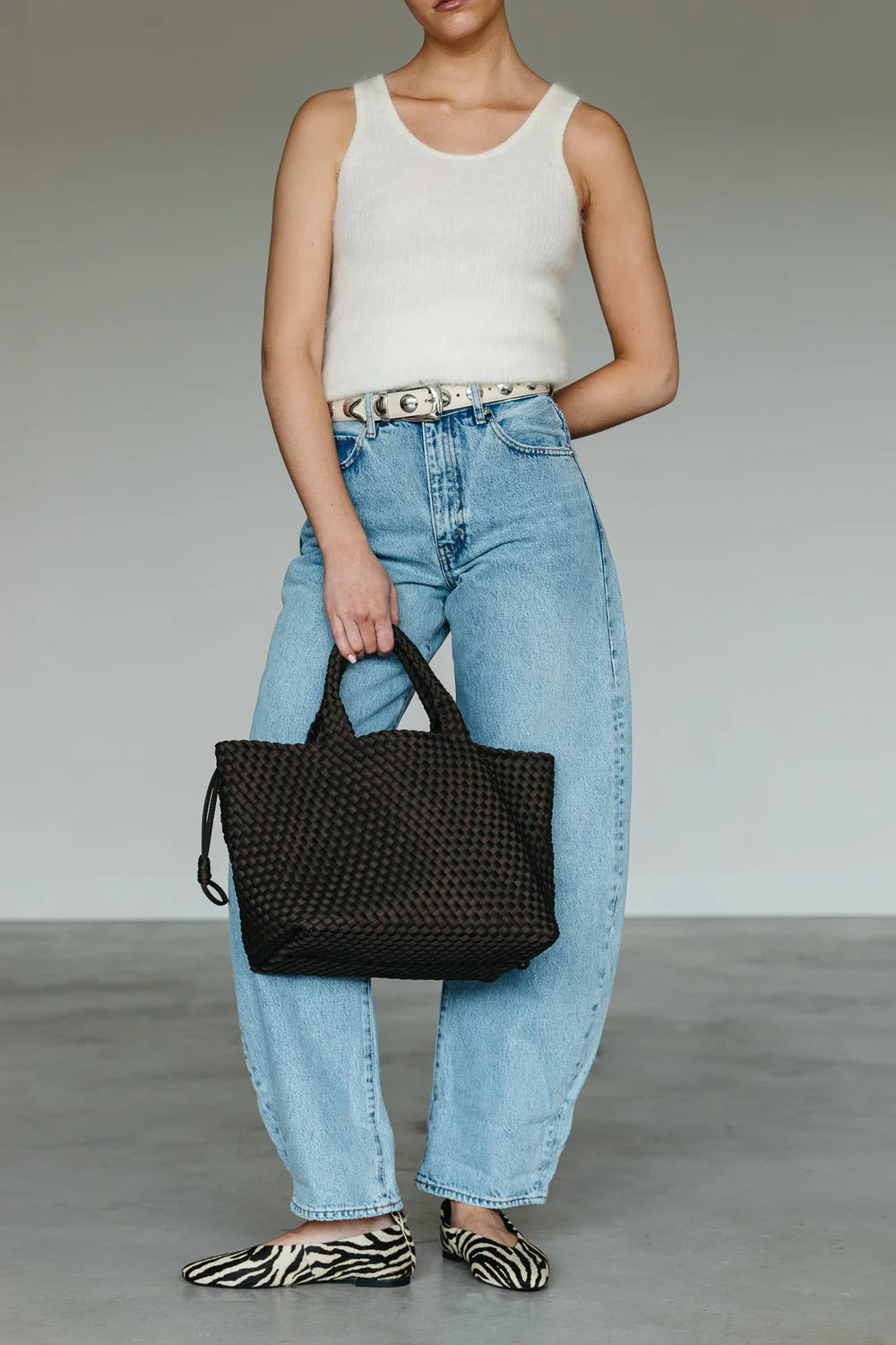 Person holding a black woven handbag wearing a white top and blue jeans on a neutral background