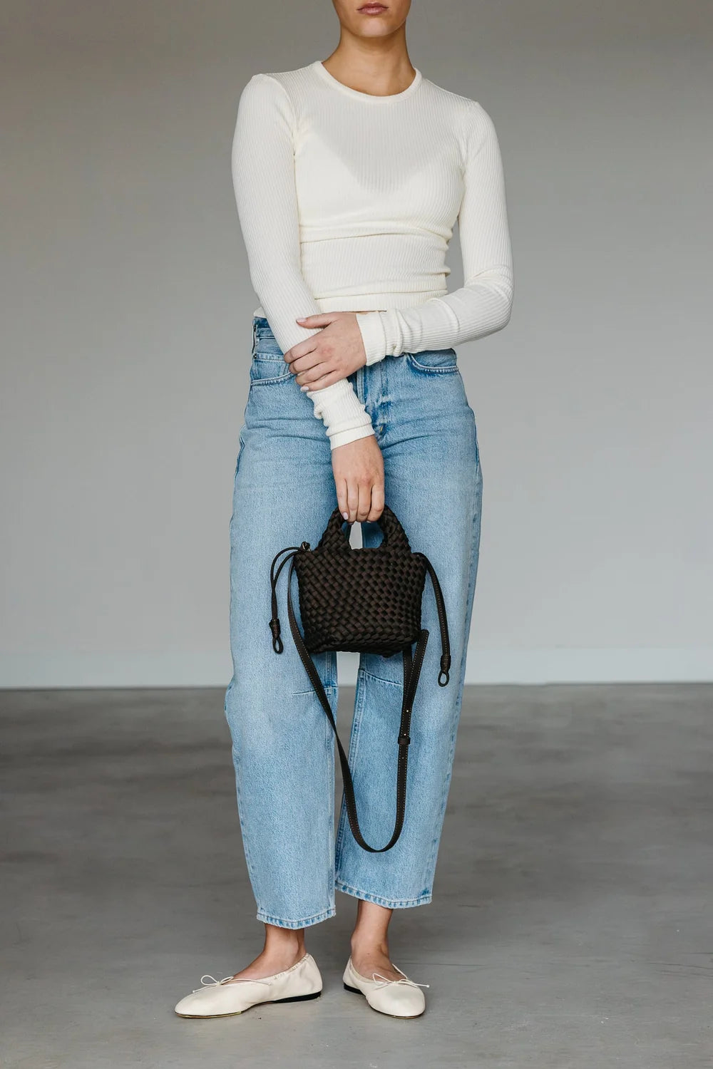 Person wearing a white long-sleeve top and blue jeans, holding a black woven handbag on a plain background