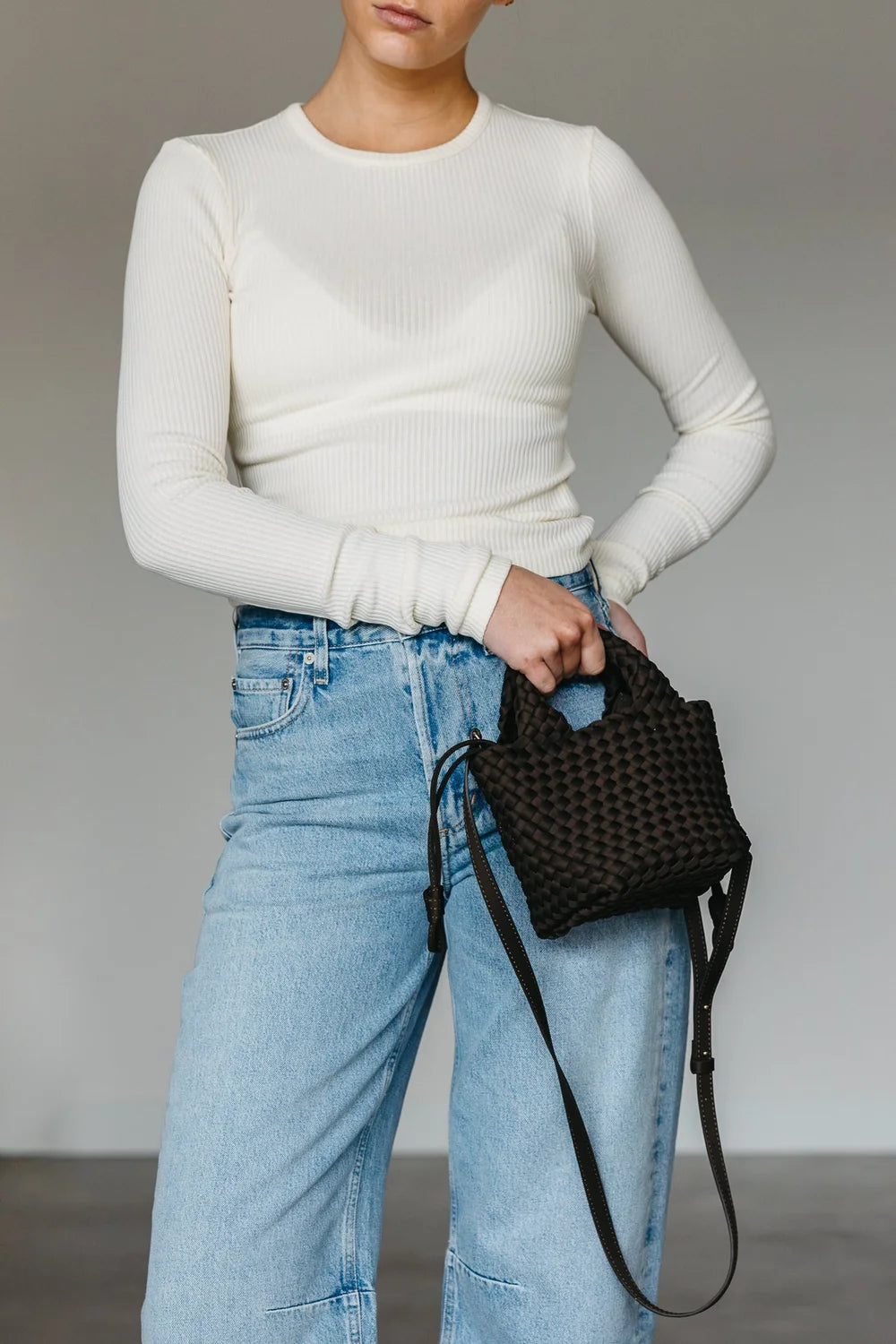 Person wearing a white long-sleeve top and blue jeans, holding a black woven handbag against a neutral background.