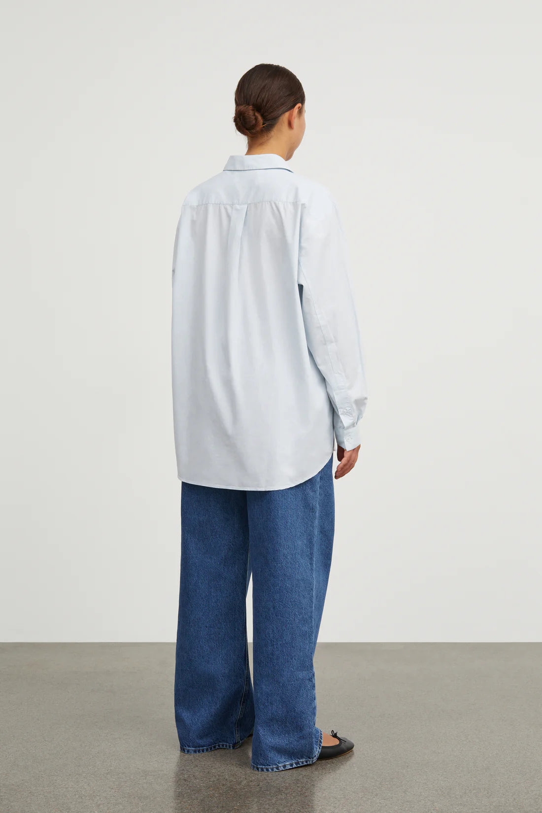 Person wearing a light blue shirt and blue jeans standing against a white wall.