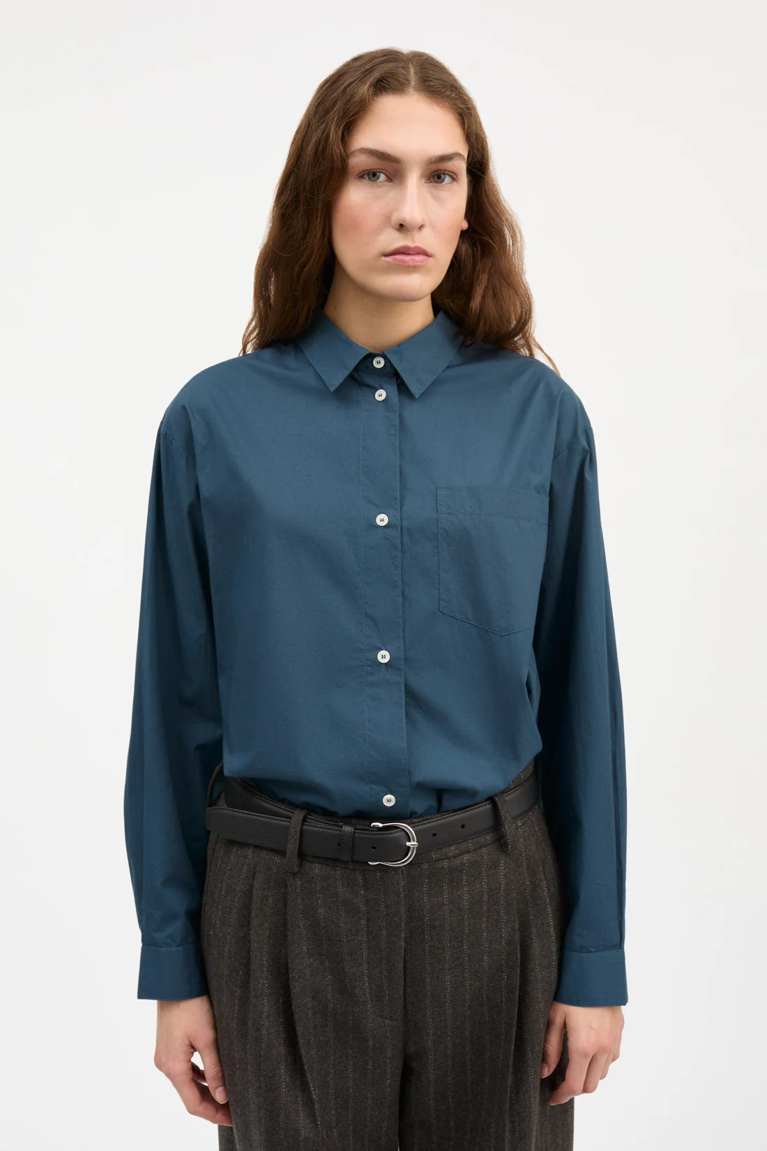 A woman wearing a slate blue long-sleeve button-down shirt tucked into dark gray pinstripe trousers with a black belt, photographed against a plain light gray studio background.