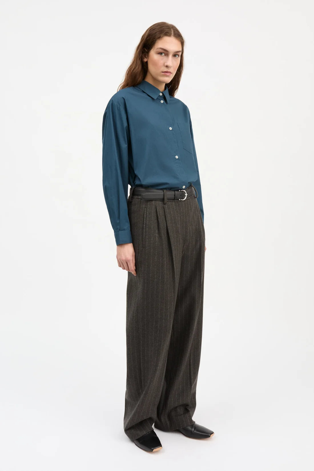 A model stands against a plain white studio background wearing a teal-blue long-sleeve button-up shirt tucked into dark charcoal gray wide-leg trousers with a belt.