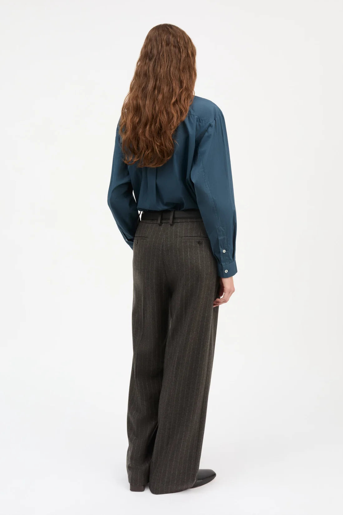 A woman with long wavy brown hair is shown from behind wearing a dark teal long-sleeve button-up shirt and dark gray pinstriped wide-leg trousers, standing against a plain white studio background.