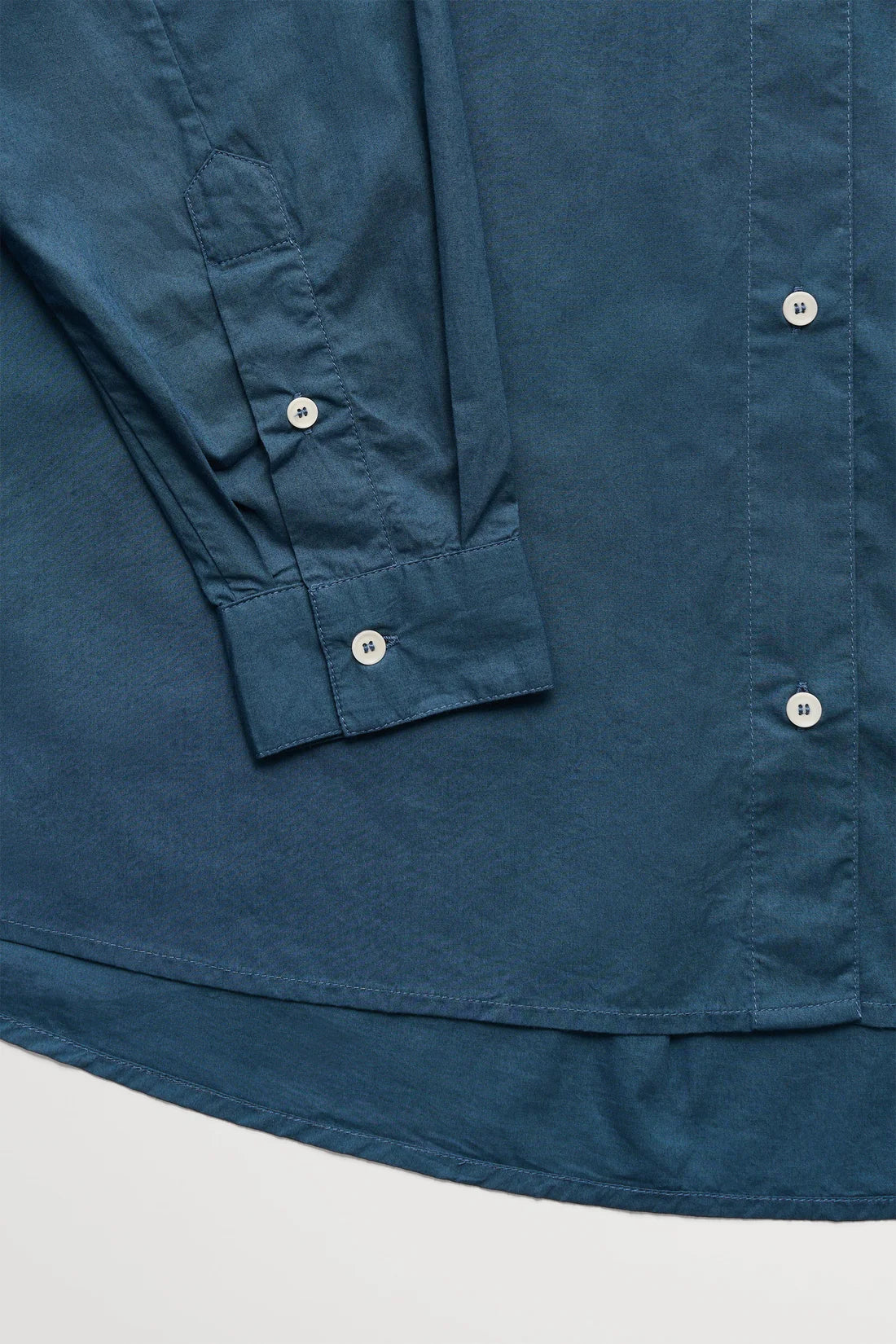 Close-up of a dark teal-blue long-sleeve button-up shirt showing the fabric texture, cuff detail, and white buttons against a light gray background.