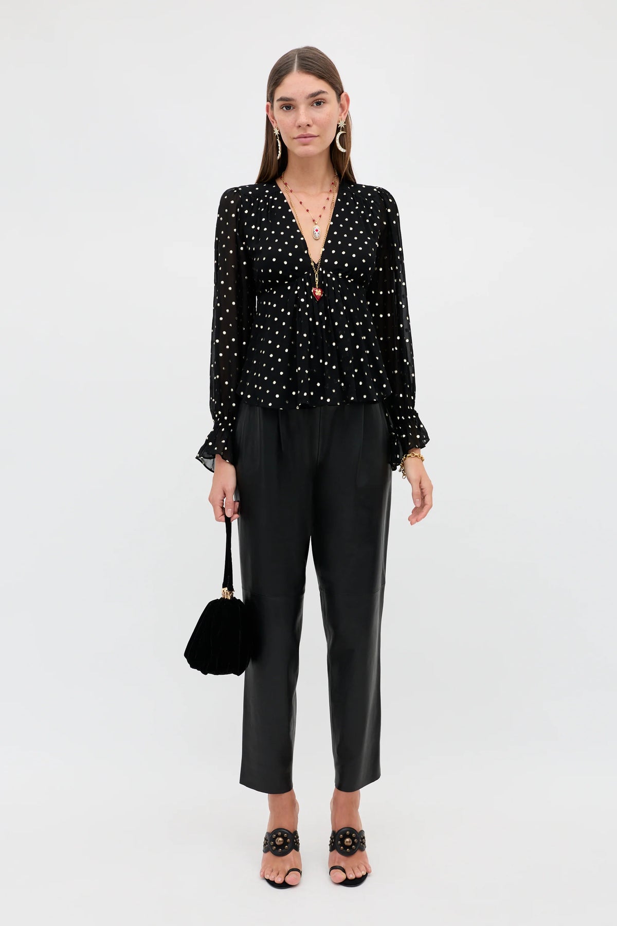 A model stands against a light gray studio background wearing a black long-sleeve blouse with white polka dots and ruffled cuffs, paired with black cropped trousers, black heeled sandals, and holding a small black handbag.