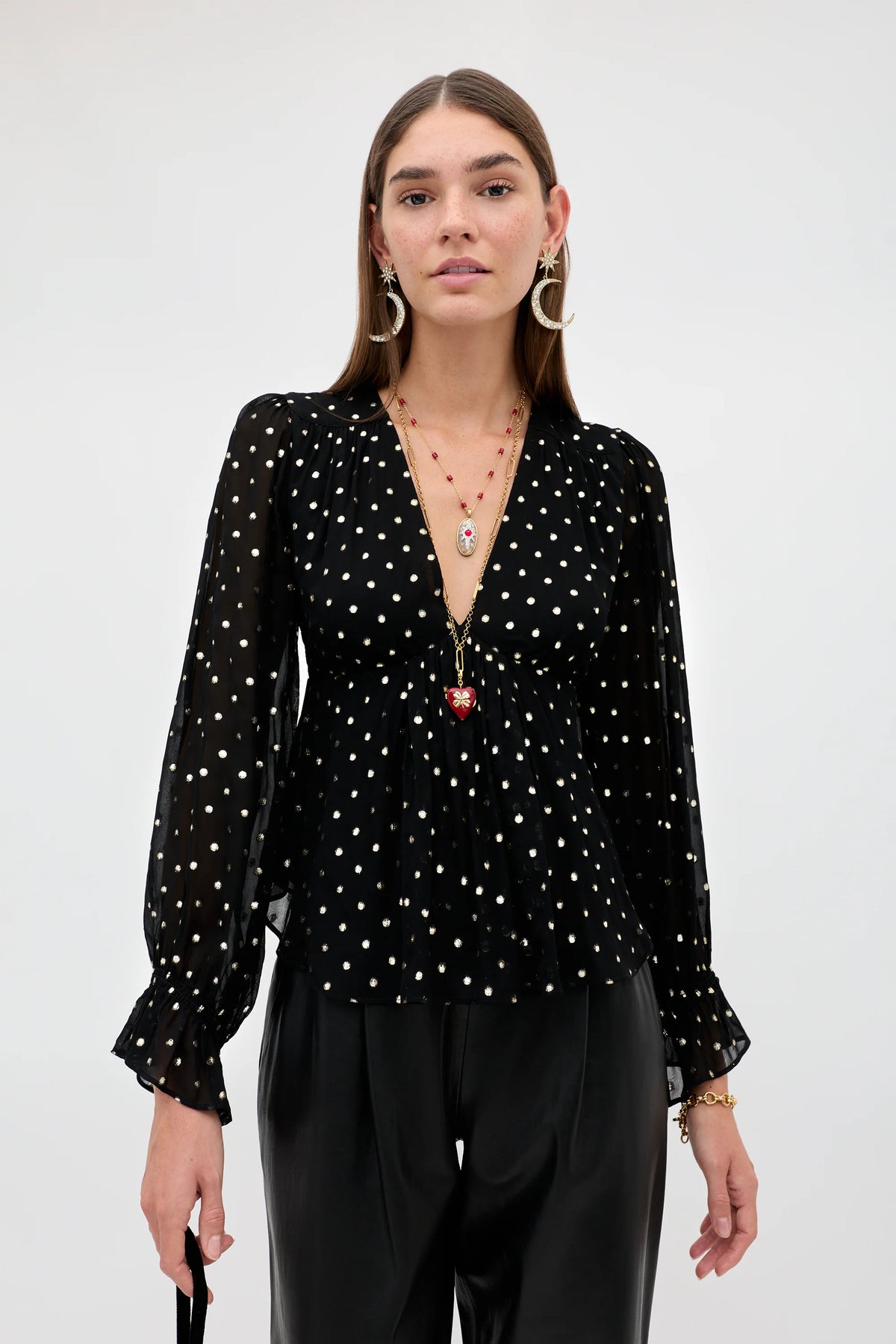 A woman modeling a black long-sleeve blouse with white polka dots, paired with black trousers, against a plain light gray studio background.