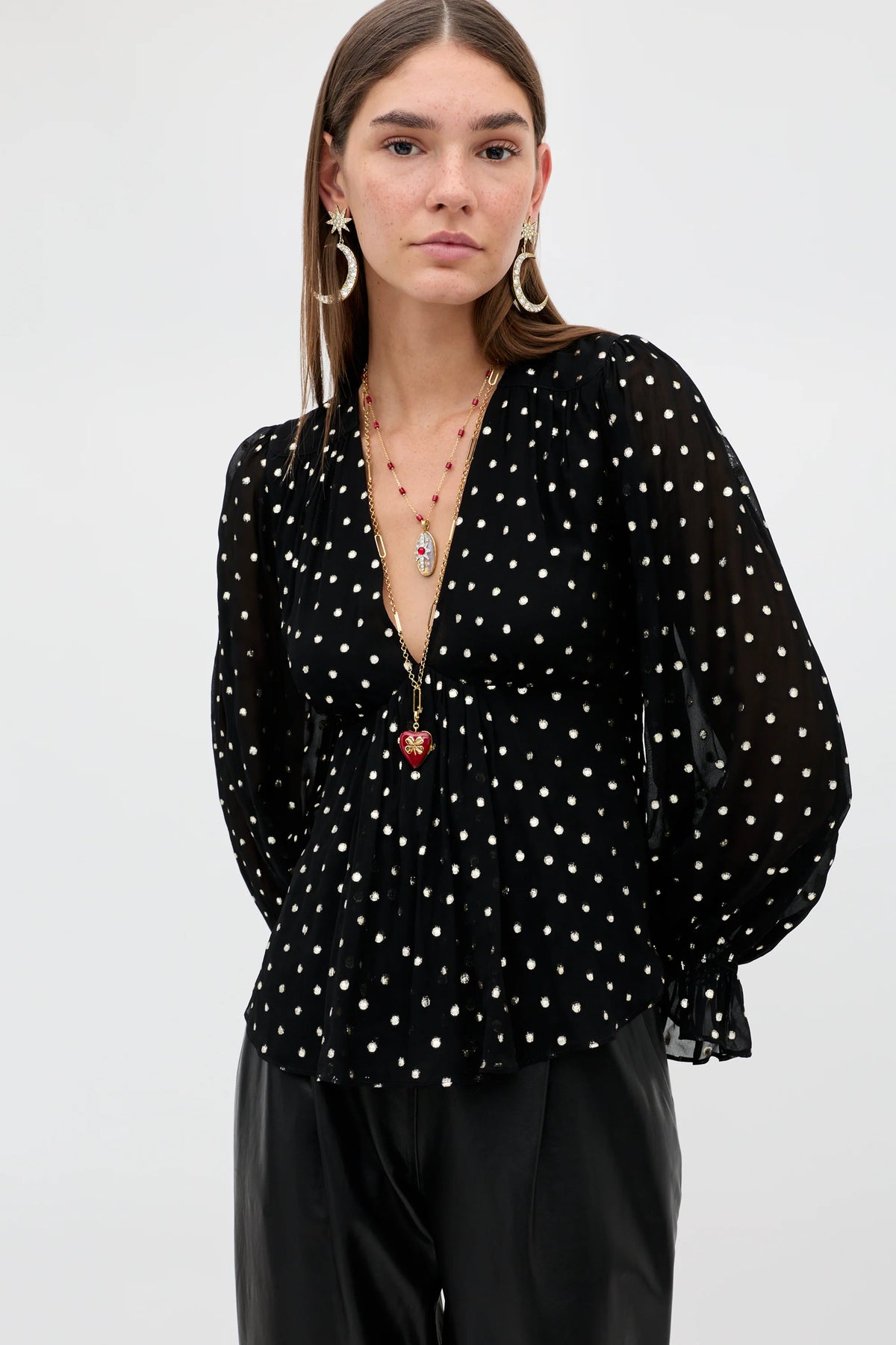 A woman modeling a black sheer long-sleeve blouse with white polka dots, paired with black trousers, against a plain light gray studio background.