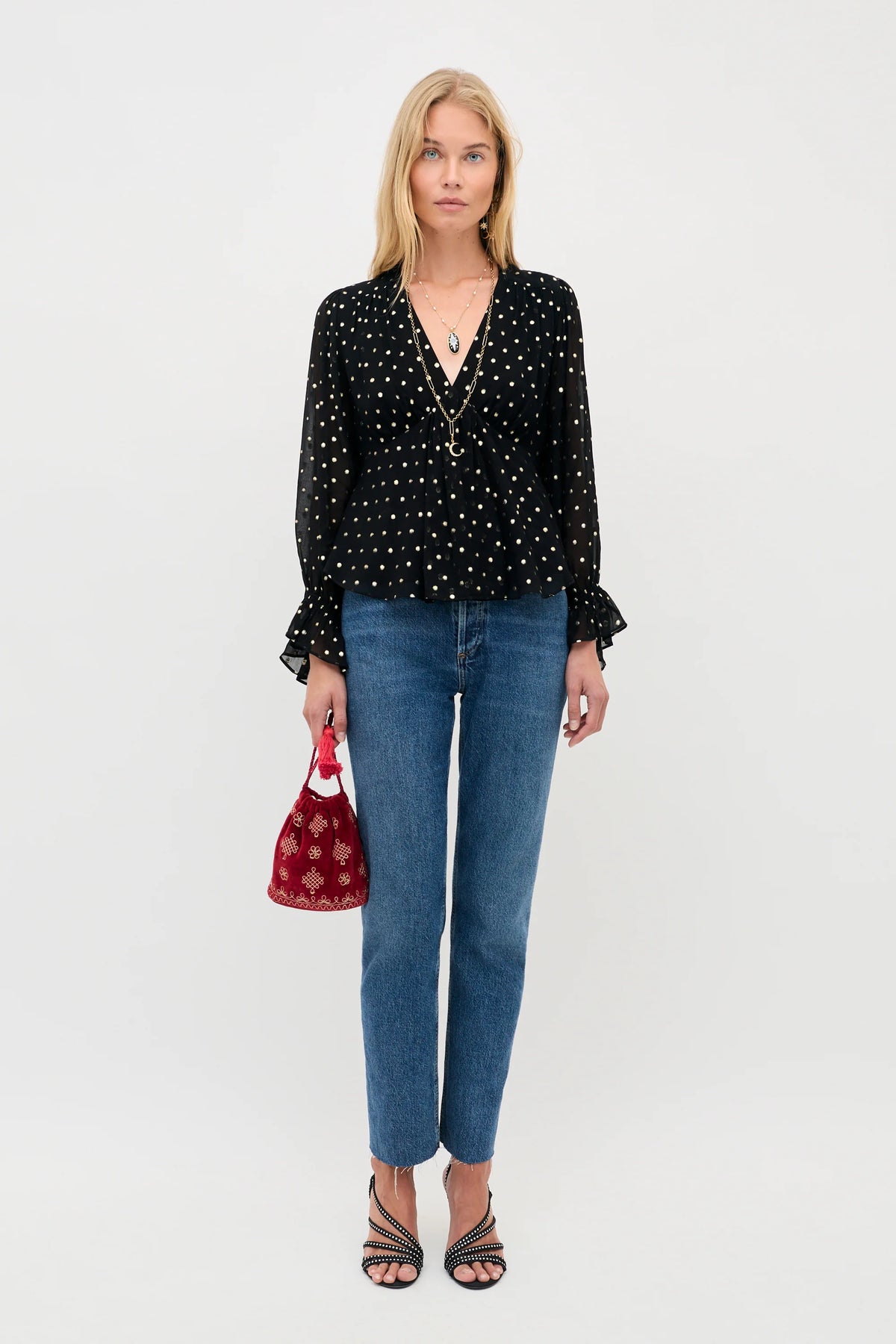 A woman stands against a light gray studio background wearing a black long-sleeve blouse with white polka dots and ruffled cuffs, blue jeans, and black strappy heels, while holding a small red patterned handbag.