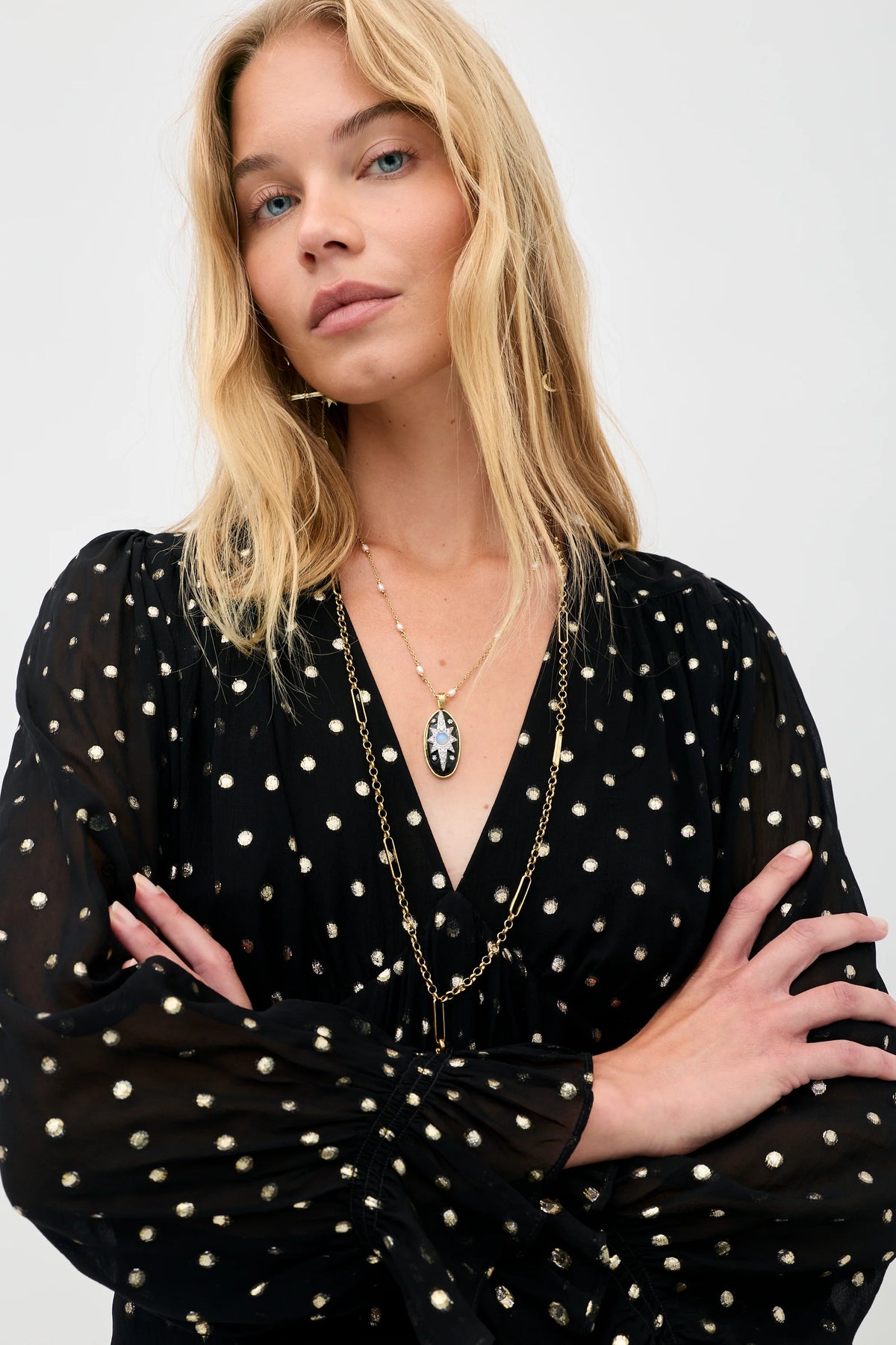 A blonde woman wearing a sheer black long-sleeve blouse with white polka dots, layered with gold necklaces, against a plain light gray background.