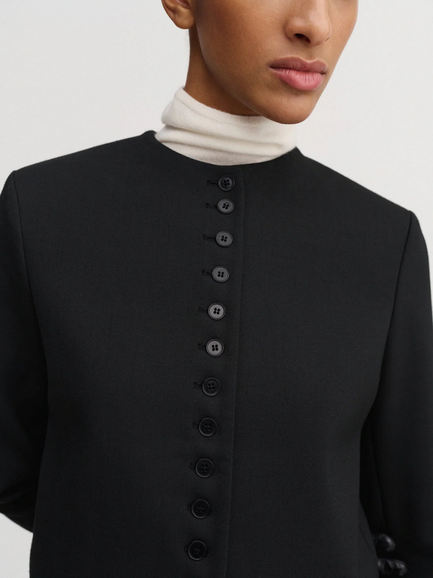 Close-up of a model wearing a black long-sleeve top with a high mock neck and a vertical row of buttons down the front, layered over a white turtleneck.