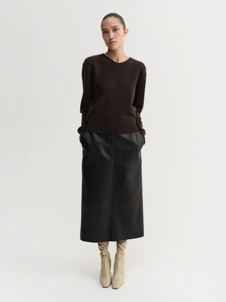 A model stands against a light gray studio background wearing a dark brown crewneck knit sweater, a black midi skirt, and beige ankle boots.