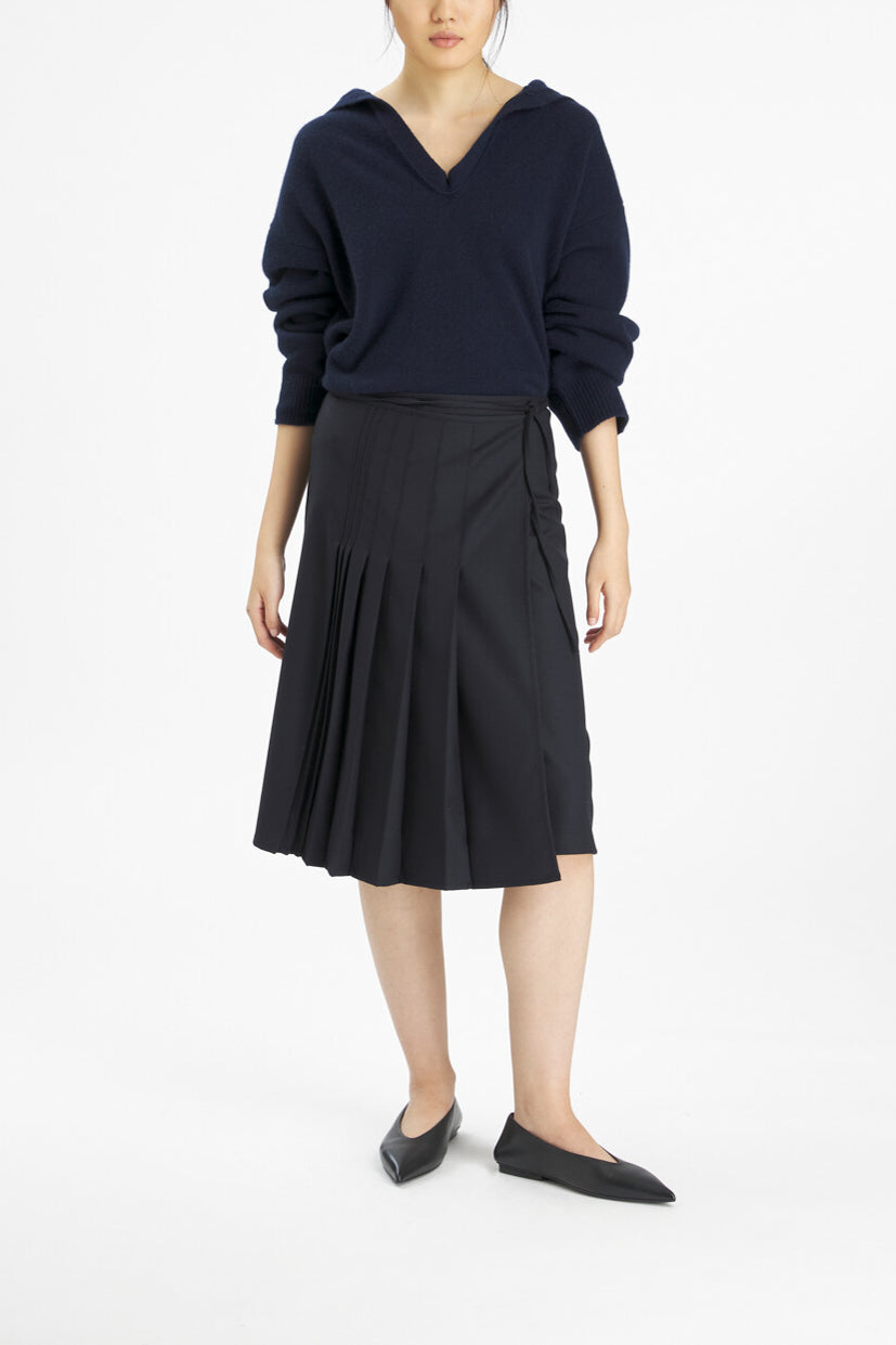 A model wearing a black long-sleeve knit top and a black pleated wrap-style midi skirt, paired with black pointed flats, against a plain white studio background.
