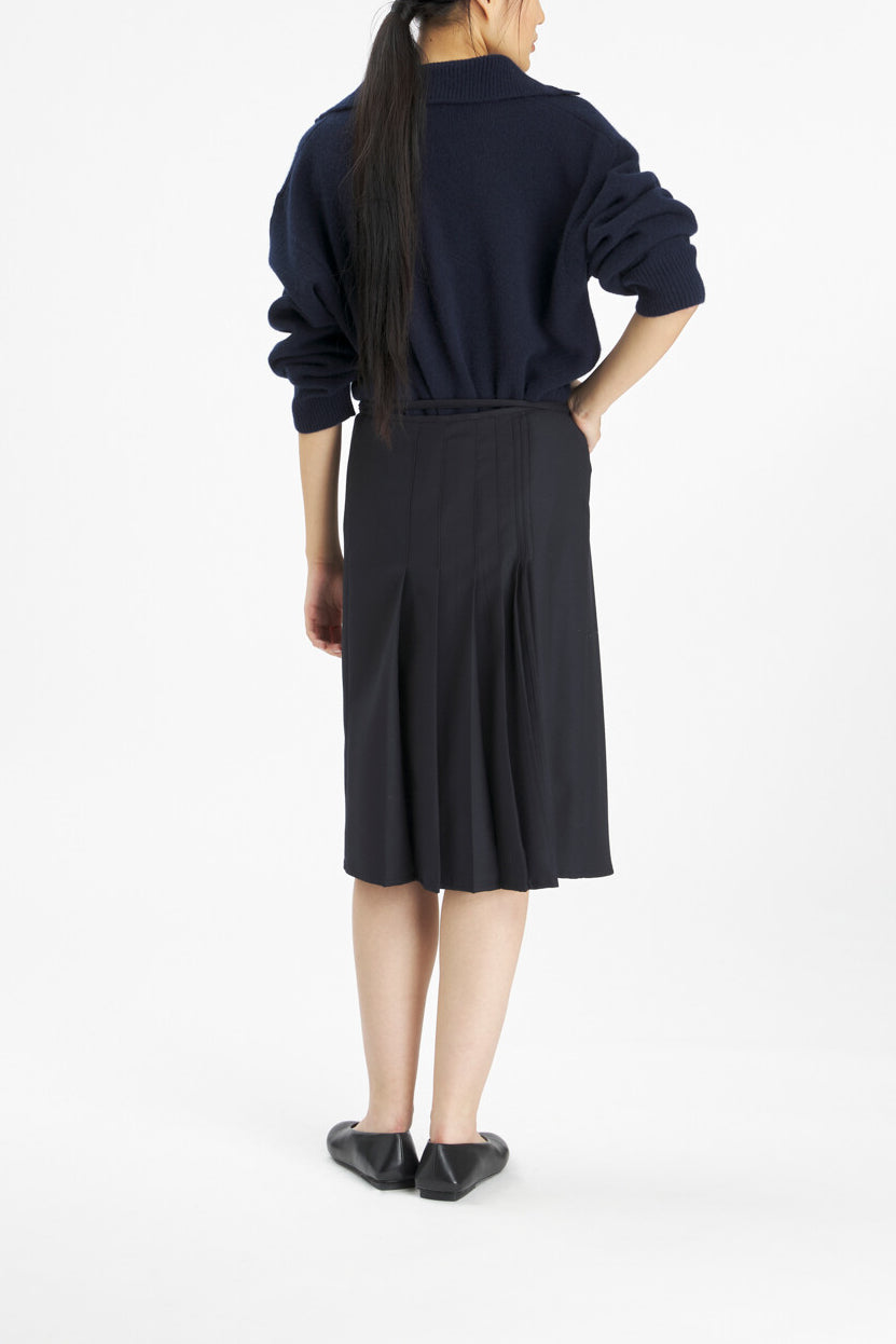 A model shown from the back wearing a dark navy blue long-sleeve collared top and a matching dark pleated midi skirt, paired with black flats, against a plain white studio background.