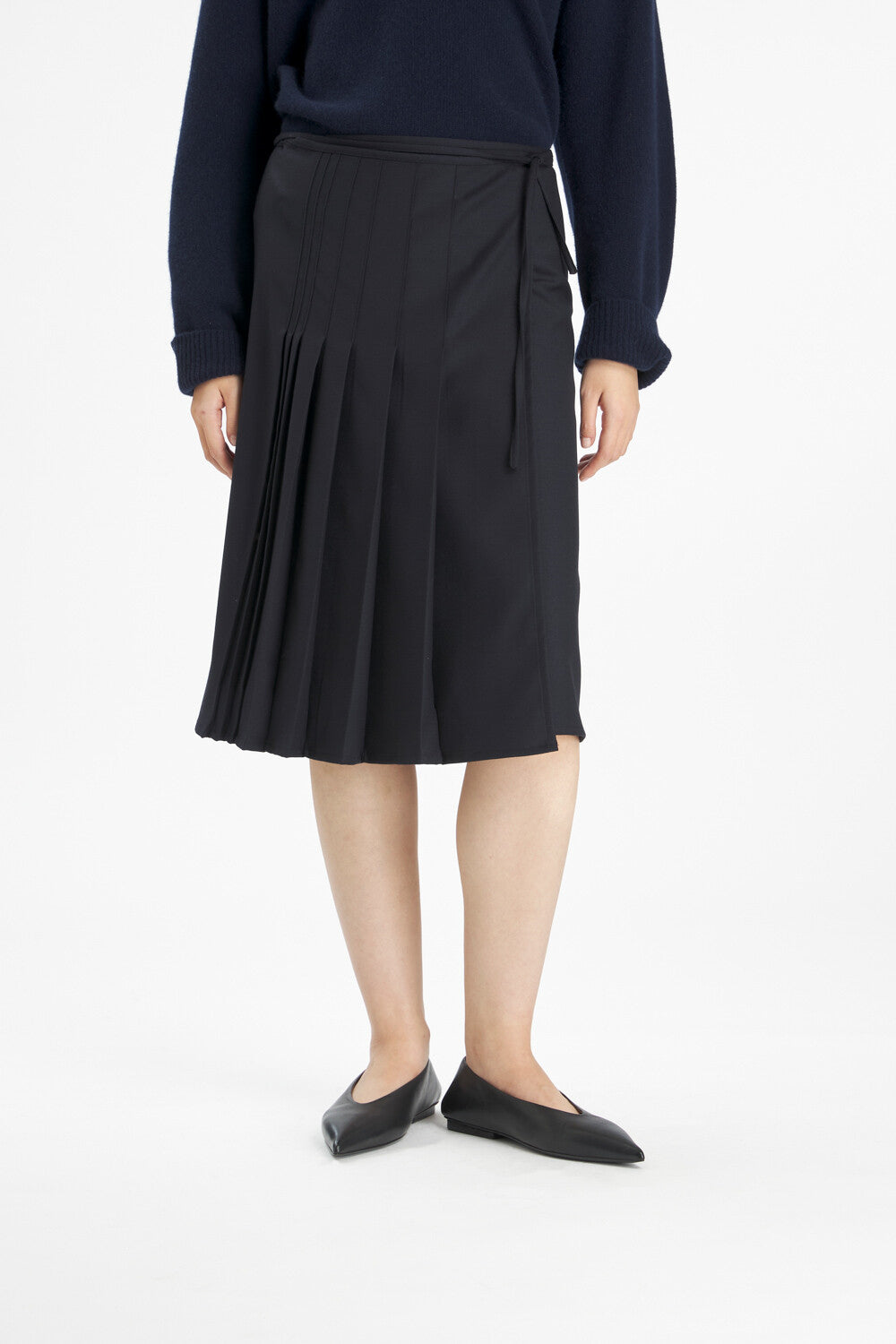 A model wearing a black pleated wrap midi skirt with a side tie, styled with a navy long-sleeve top and black pointed flats against a white studio background.