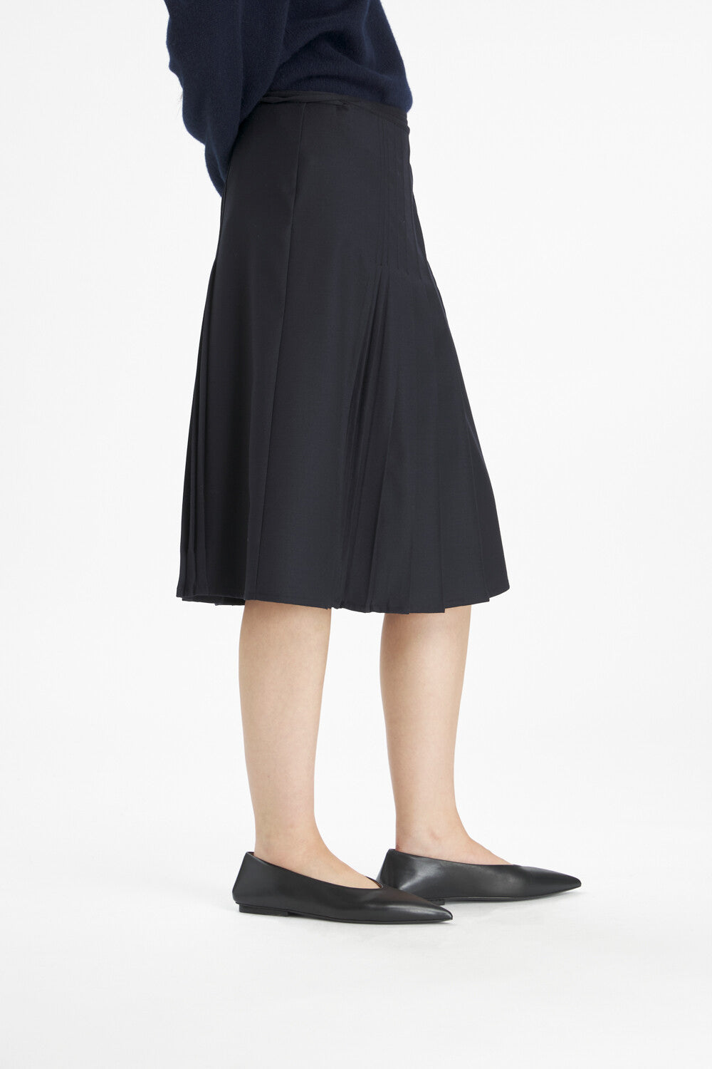 A person wearing a black pleated midi skirt with a navy top and black pointed flats, shown from the waist down against a plain white studio background.
