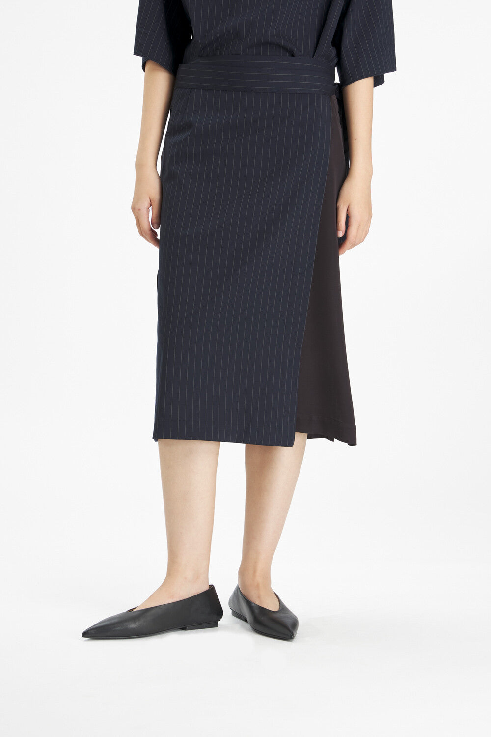 A model wearing a dark navy blue pinstriped midi skirt with a side slit and black pointed flats, shown from the waist down on a white studio background.