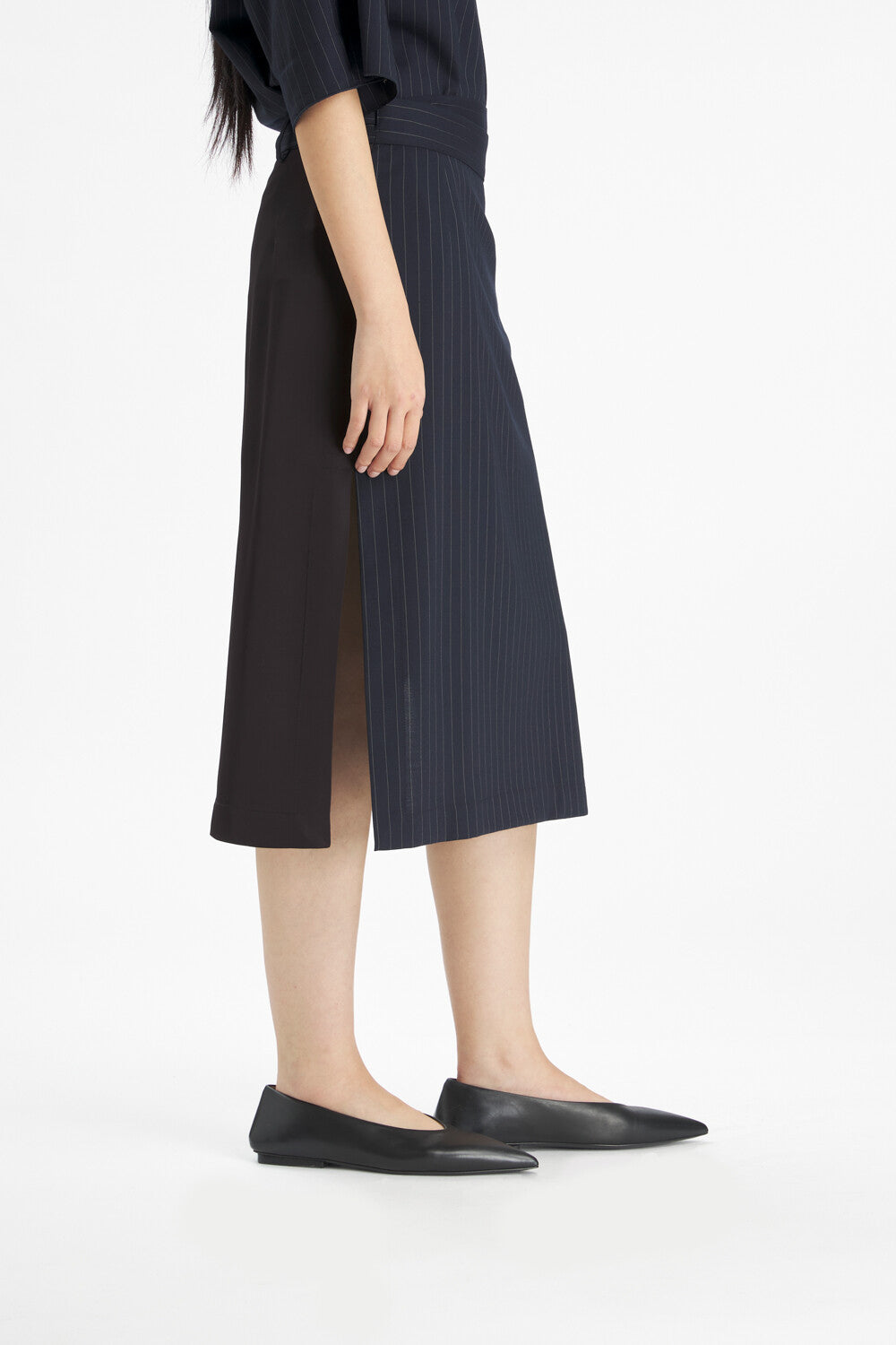 A model wearing a dark navy blue pleated midi skirt with a side slit, paired with black pointed-toe flats, shown from the waist down against a plain white studio background.