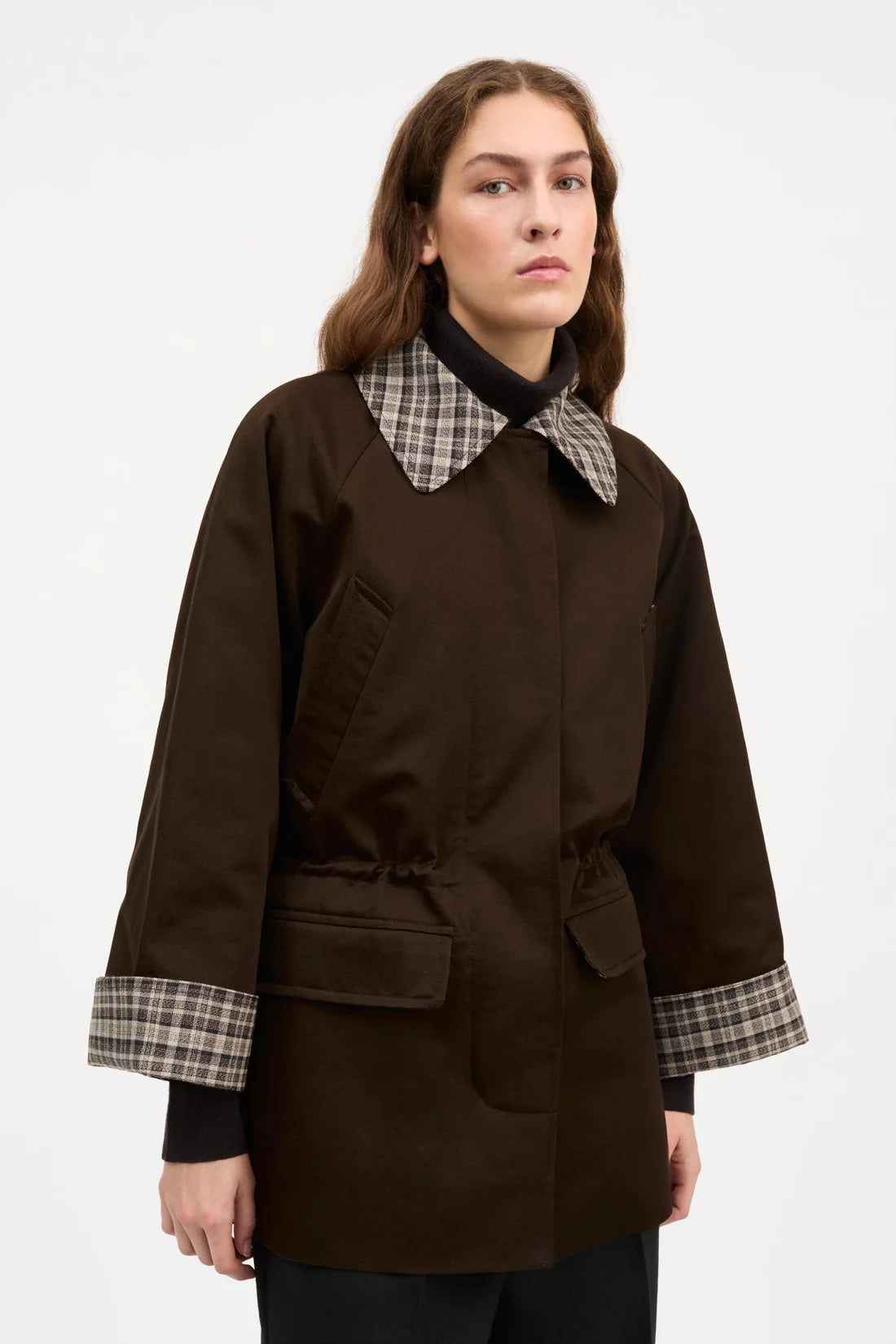 A model wearing a dark brown utility-style jacket with a contrasting plaid collar and matching plaid turned-back cuffs, shown against a plain light gray studio background.