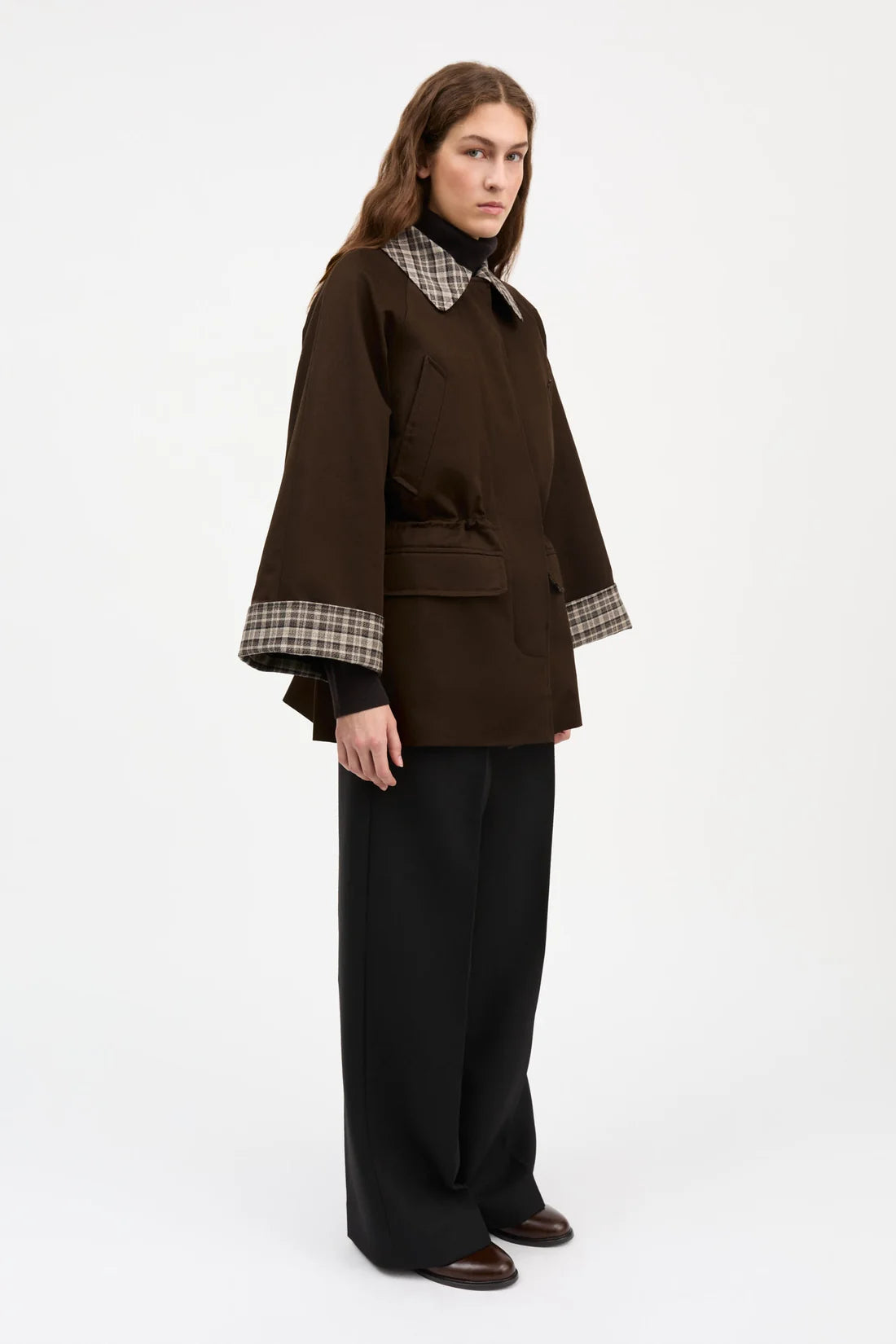 A model stands against a plain white studio background wearing a dark brown cropped jacket with a plaid collar and cuffs, paired with black wide-leg trousers and brown shoes.