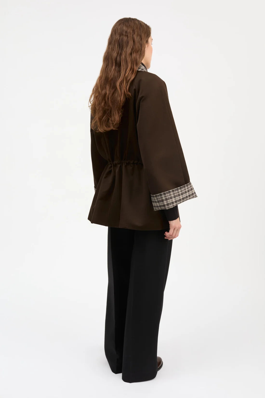 A woman shown from the back wearing a dark brown jacket with a plaid cuff lining and black wide-leg trousers, photographed against a plain white studio background.