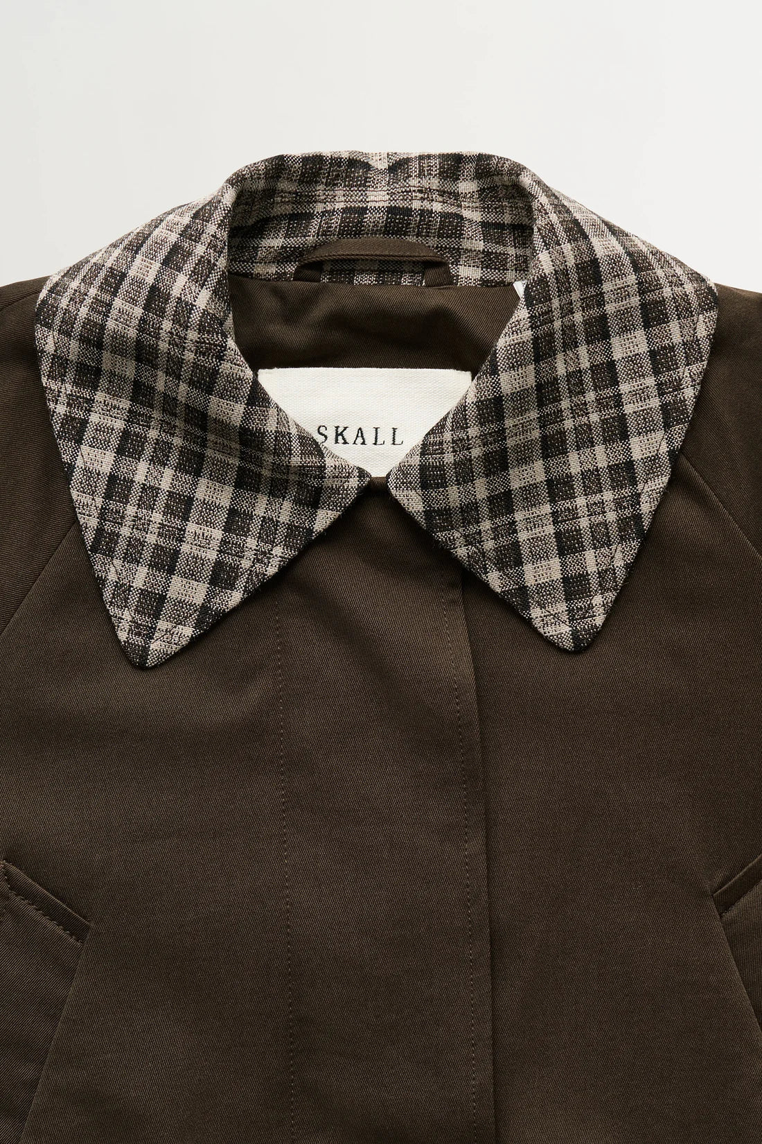 Close-up of a dark brown coat with a contrasting plaid collar in black, white, and gray, featuring a visible SKALL brand label.