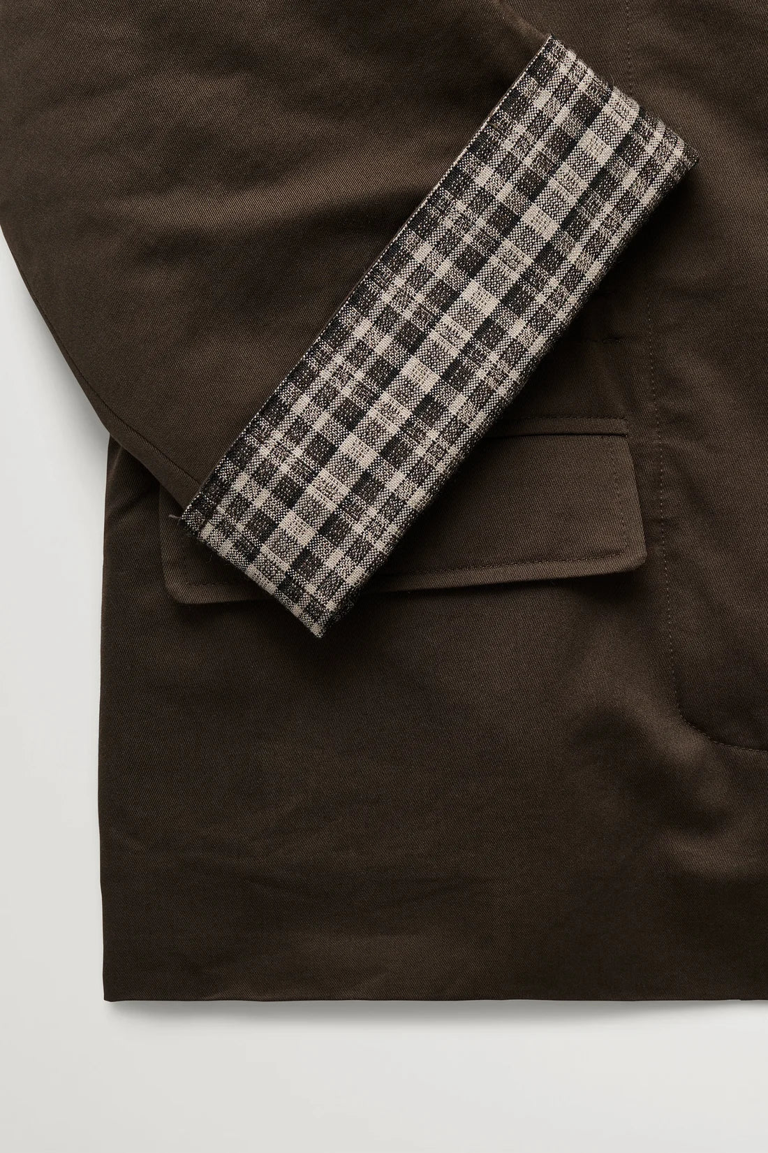 Close-up of a dark brown jacket with a folded cuff showing a black, gray, and beige plaid lining, set against a plain light gray background.