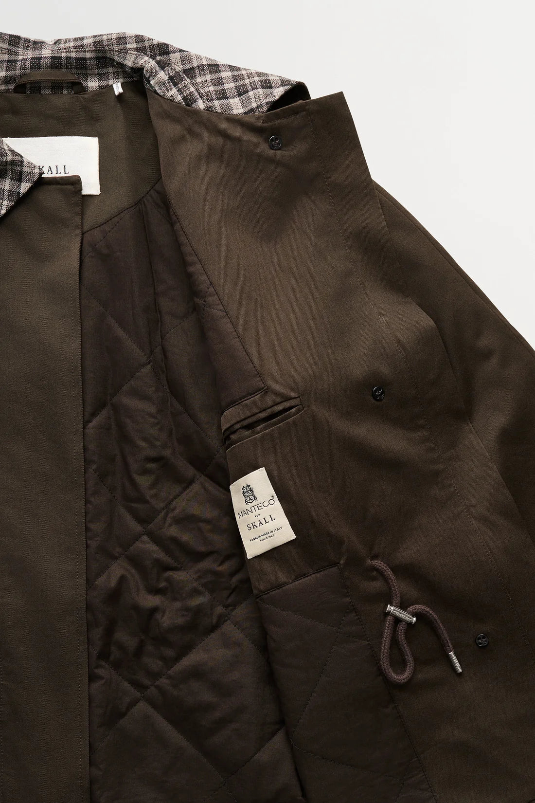 Close-up of a dark brown quilted jacket by The Rough Trade, showing the interior lining, a plaid collar, and a sewn-on brand label.